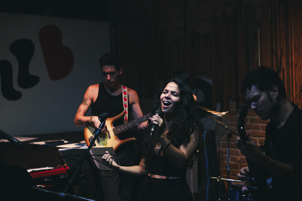 A musical band performs on stage, with a female singer passionately singing into a microphone, a male guitarist on her right, and a male bassist on her left, all in a dimly lit venue with brick walls.
