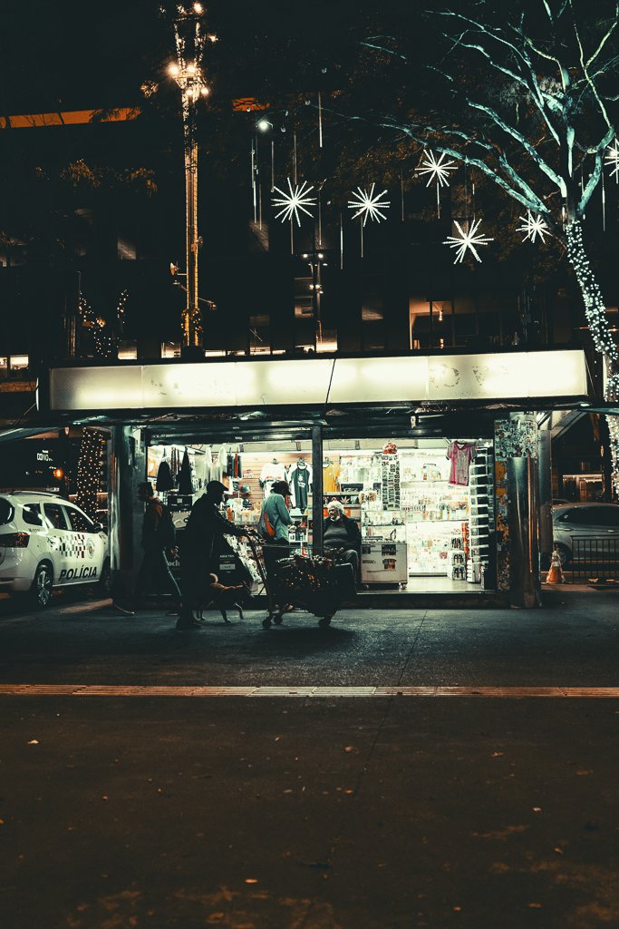A small illuminated kiosk or convenience store at night with people shopping outside. Christmas lights and star-shaped decorations hang from a tree and the building. A police car is parked nearby.