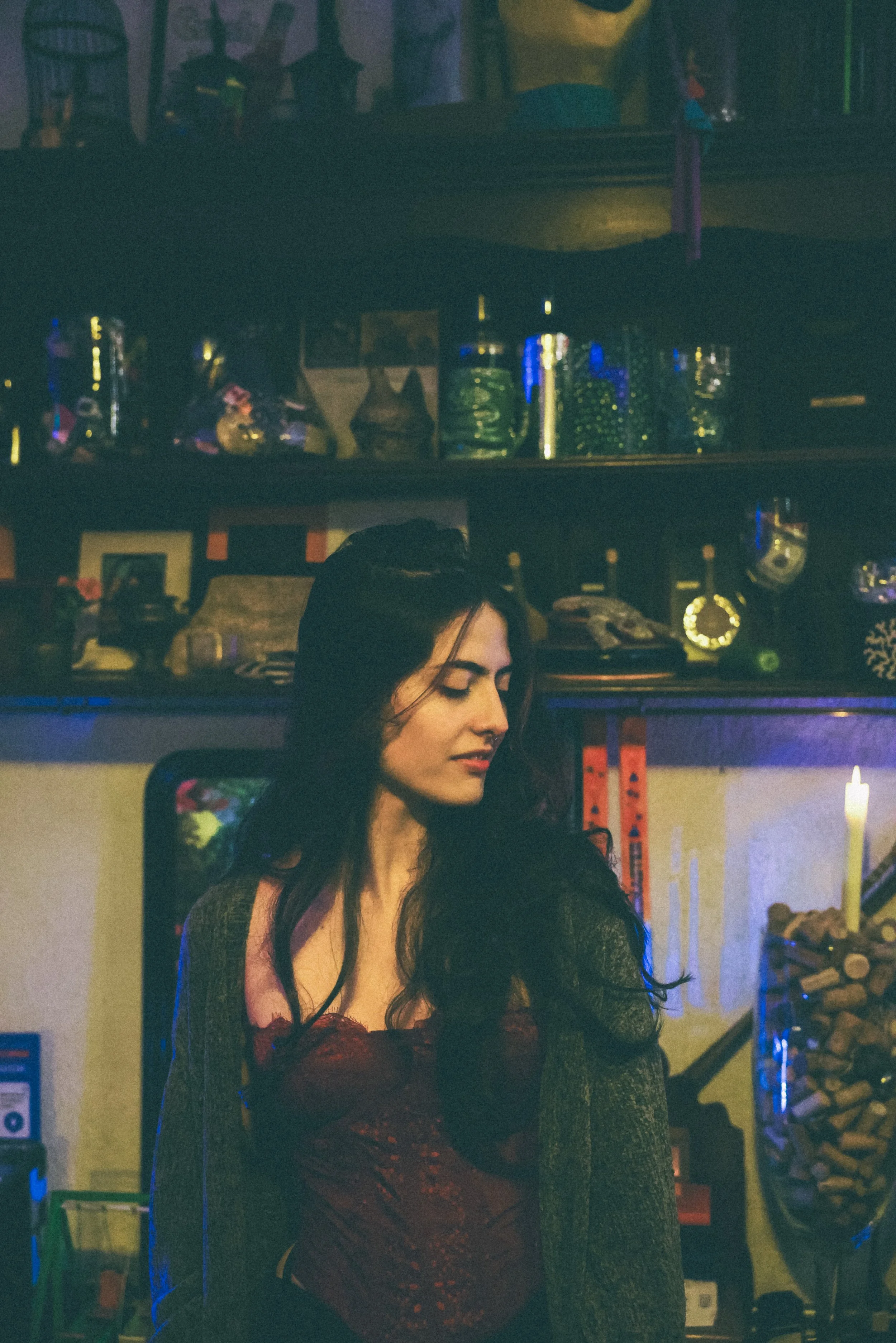 A woman with dark, wavy hair wearing a dark gray cardigan over a red, strapless top, standing in a dimly lit room with shelves filled with various jars, bottles, and decorative items.