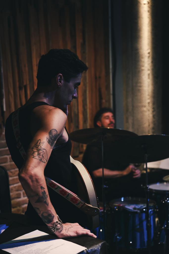A woman with tattoos on her arm playing an acoustic guitar during a performance, with a drummer in the background in a dimly lit indoor venue.