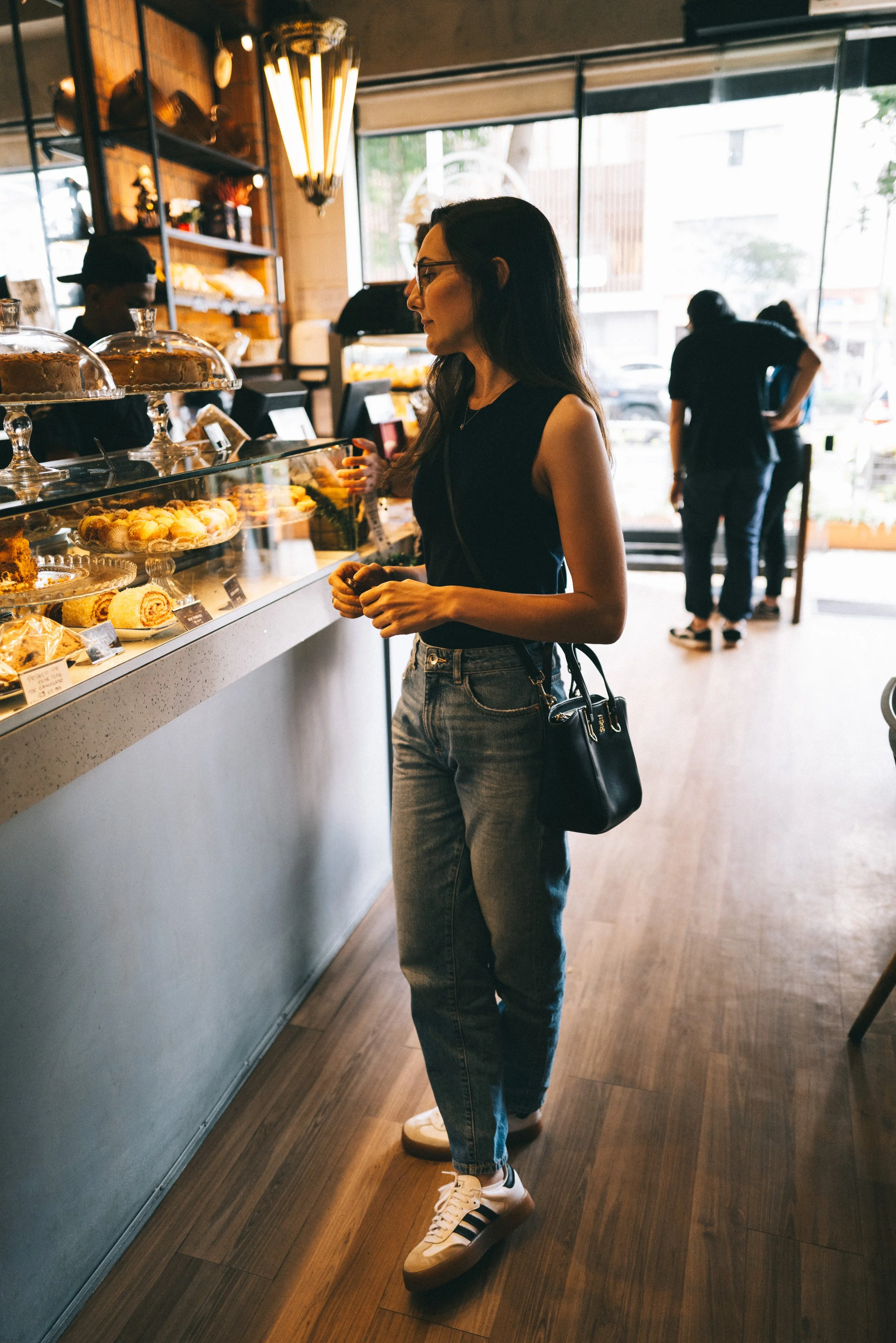 A woman with glasses, wearing a black sleeveless top, jeans, and Adidas sneakers, stands inside a bakery, looking at pastries displayed in a glass case.