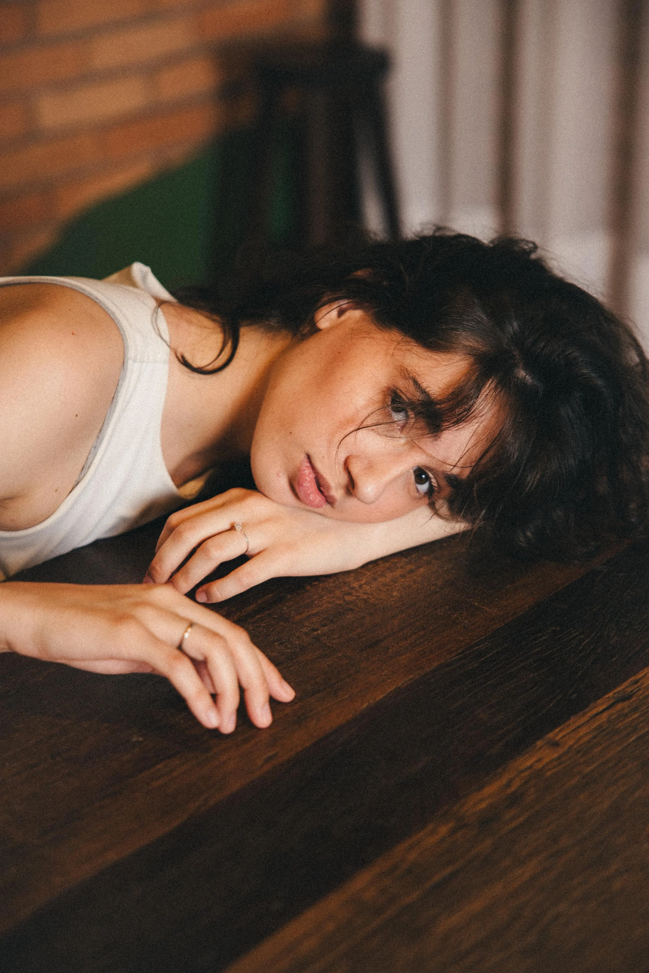 A woman with dark wavy hair lying on a wooden table, looking into the camera with a relaxed expression.