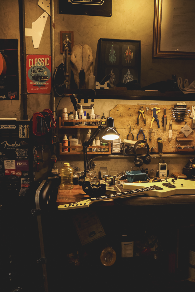 A guitar workshop with a guitar on the workbench, tools hanging on a wooden board, bottles and supplies on shelves, and various framed artwork and posters on the walls.