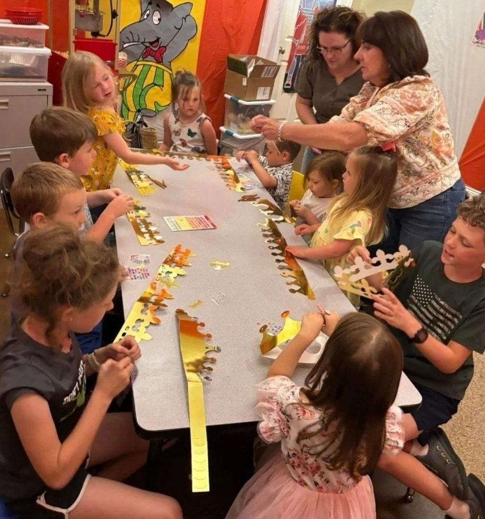 A group of children and two adult women sitting around a long table, working on crafting paper crown decorations with gold, orange, and white colors in a colorful room with cartoon character wall art.