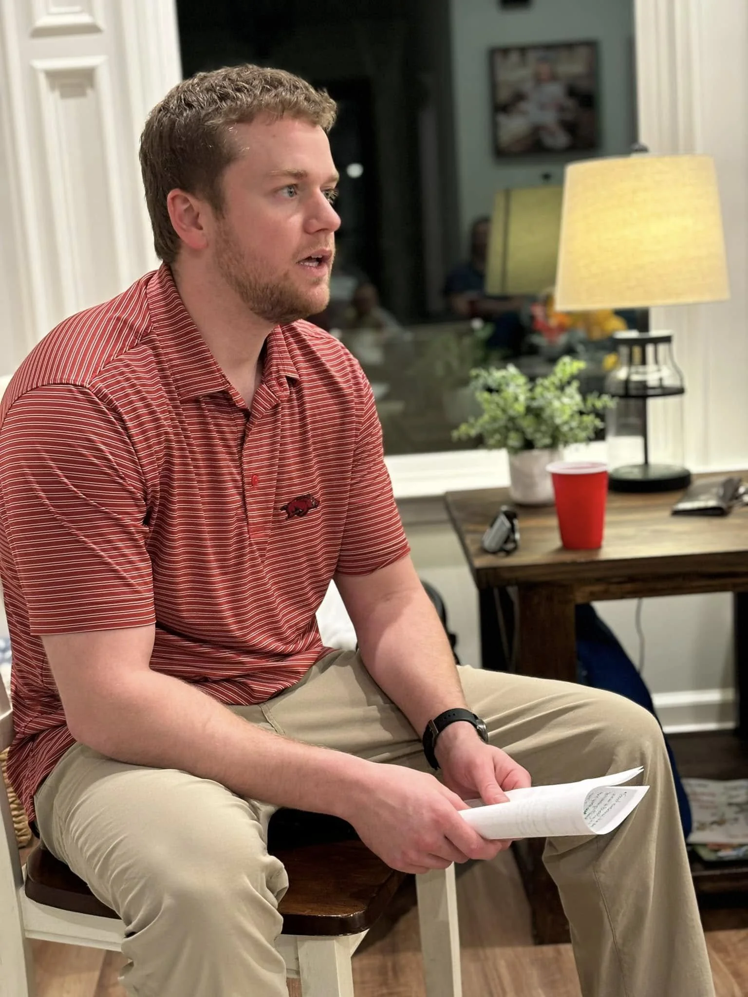 A young man with light brown hair and a beard, wearing a red striped polo shirt and khaki pants, sitting and holding papers in his hands. He appears to be speaking or reacting to something. The background shows a cozy indoor setting with a table, a red cup, a potted plant, a lamp, and a window.