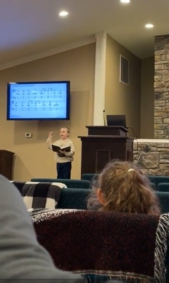 Young boy standing at a lectern in a church or classroom, holding a book and raising his hand while speaking or reciting, with a large digital screen displaying musical notes in the background, and people seated in front watching.