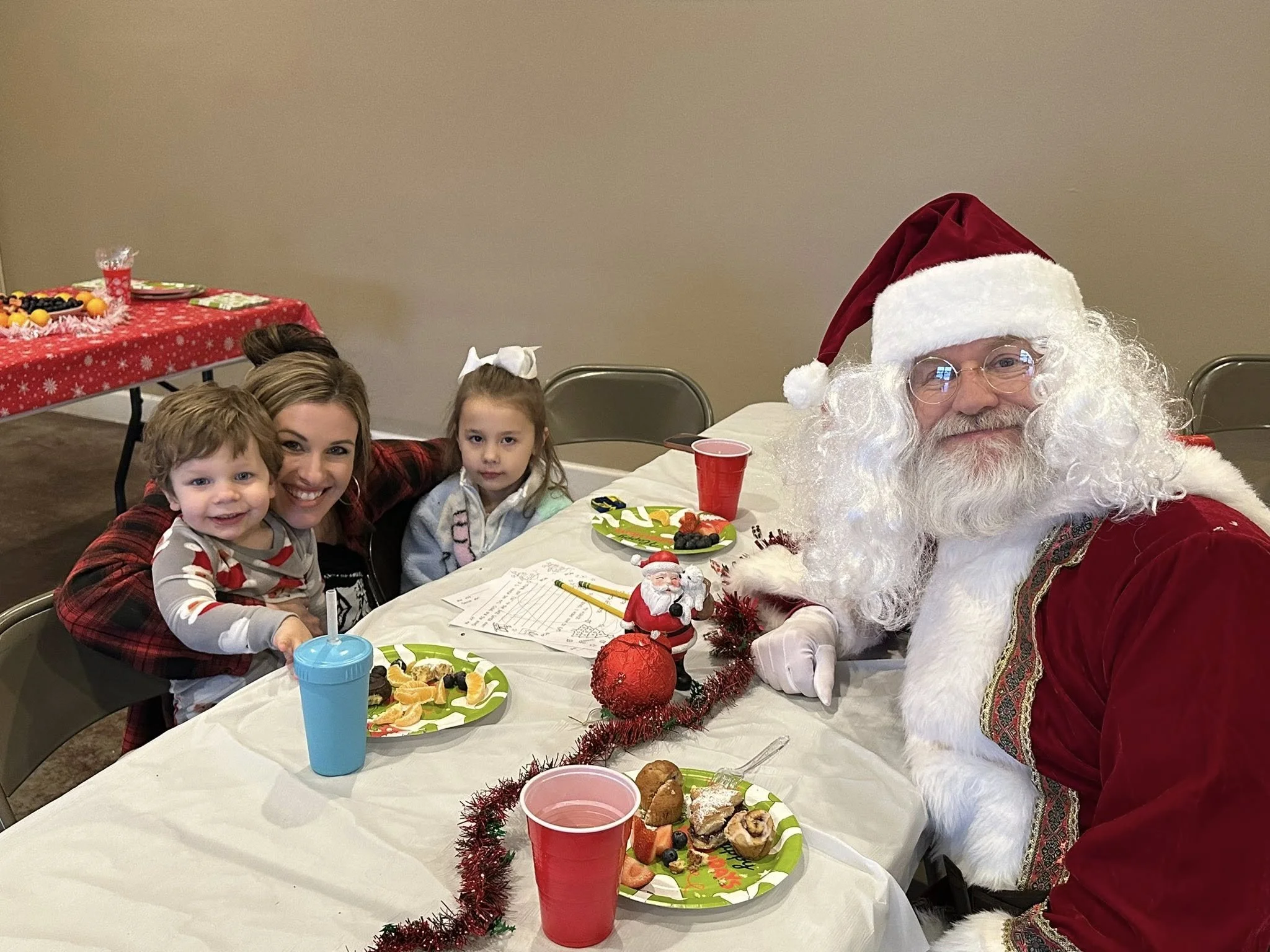 A man dressed as Santa Claus sitting at a table with two children and a woman. The woman and children are smiling. The table has plates with snacks and a small Santa figurine. In the background is another table with snacks.