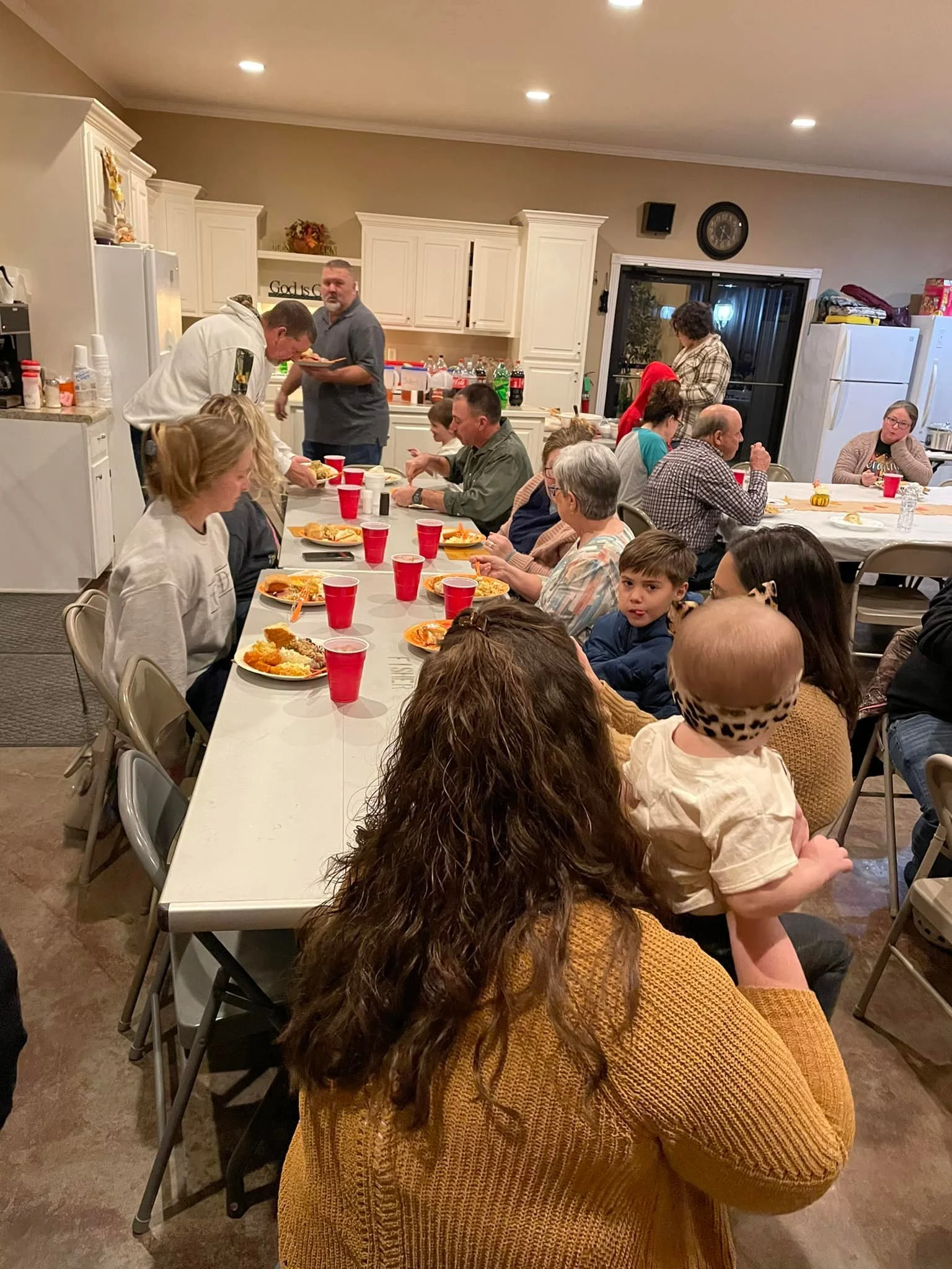 People gathered around a long table with plates of food, in a kitchen with white cabinets, during a family or social gathering.