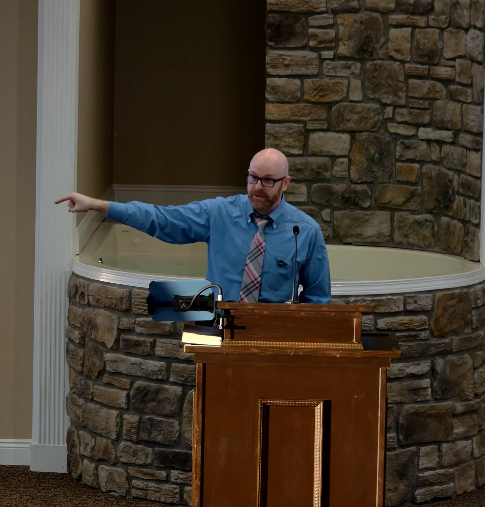A man with glasses, a bald head, and a beard is standing at a wooden podium, pointing to the left. He is wearing a blue shirt and a plaid tie. Behind him, there is a stone wall and a small indoor hot tub or jacuzzi. A microphone and a laptop are on the podium.