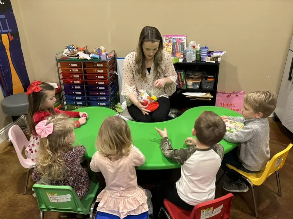 A woman leading a group of five young children in a classroom activity at a green table.