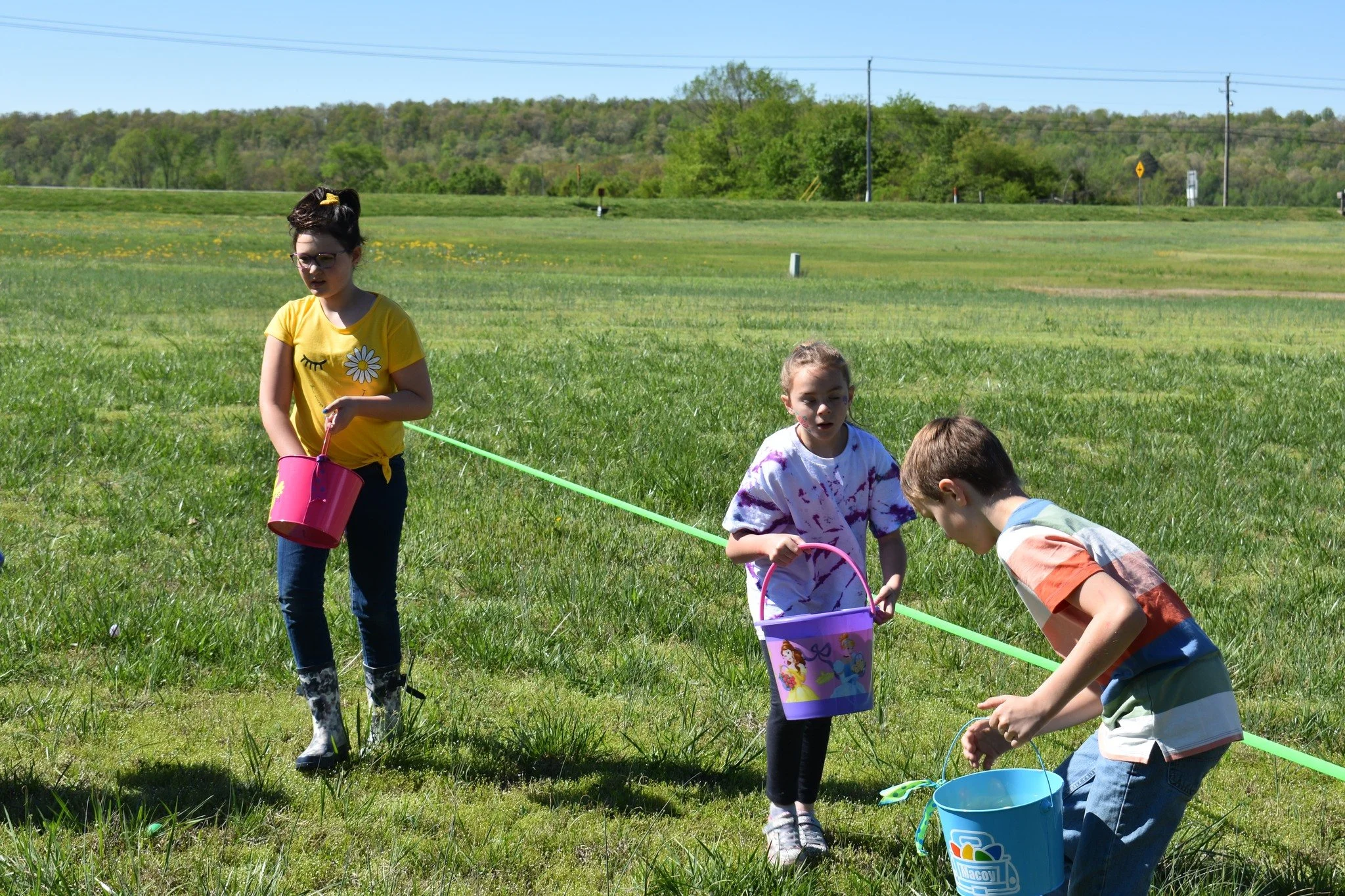 Three children outdoors in a grassy field participate in an egg and spoon race. They each hold buckets and are balancing eggs on spoons, with a green line indicating the race track.