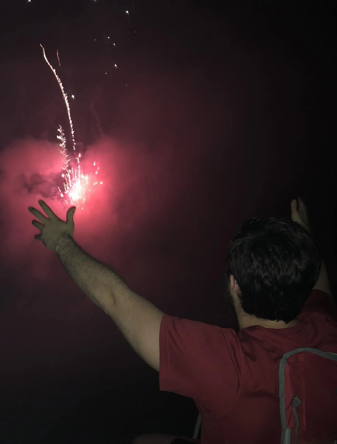 Person with dark hair and a red shirt reaching out towards fireworks in the night sky.