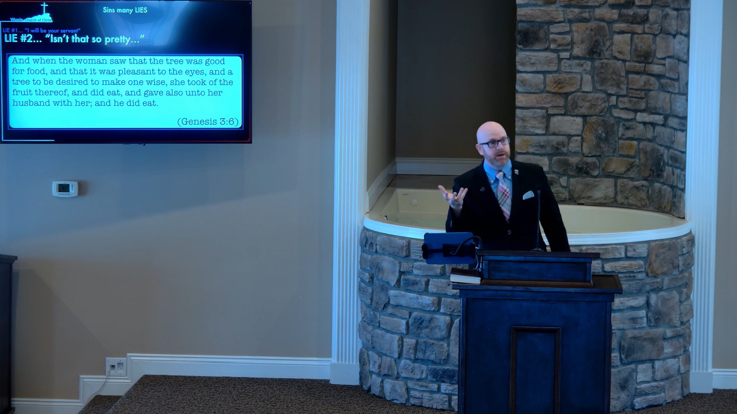 A man in a suit and glasses standing behind a wooden pulpit, gesturing with his hand, in front of a stone wall. To his left, a large screen displays a Bible verse from Genesis 3:6 with a blue background.