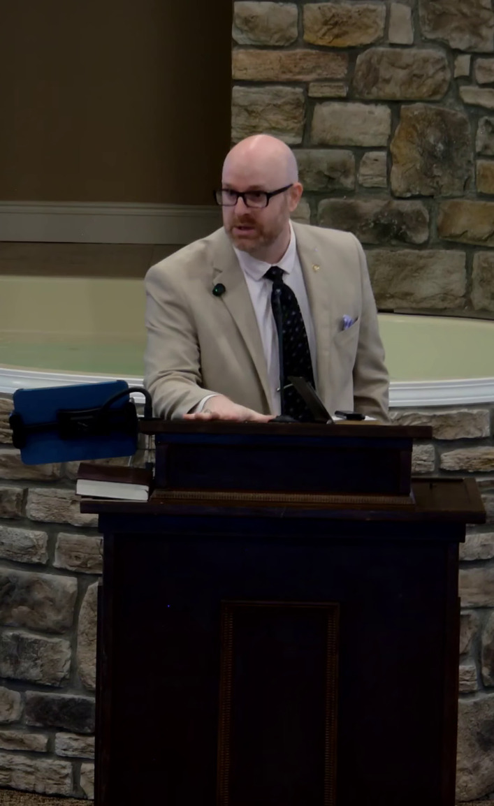 A man with glasses and a beard, wearing a beige suit and patterned tie, standing at a podium with books and electronic devices, in front of a stone wall.