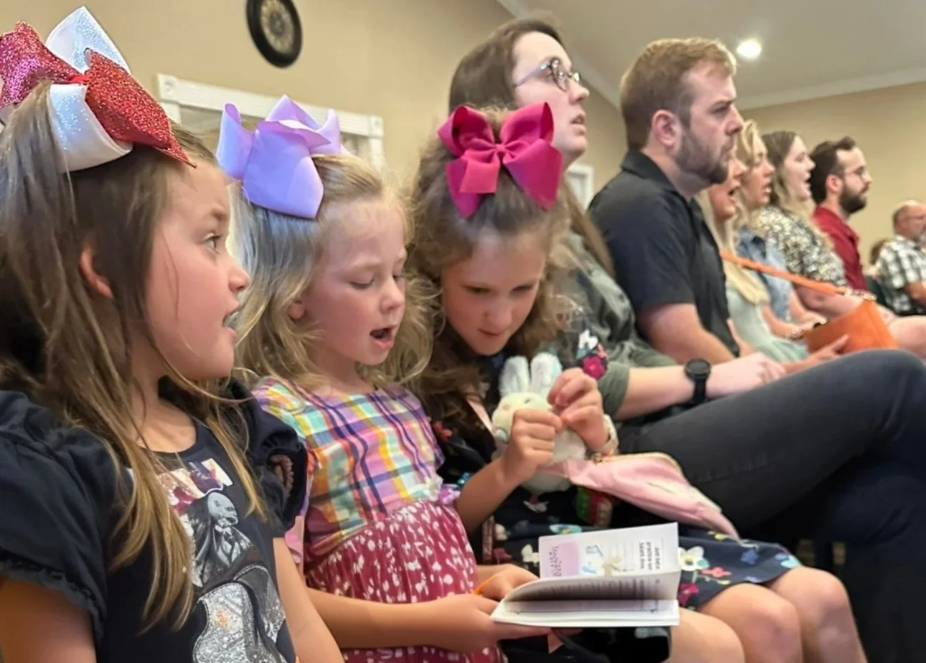 Children and adults sitting in an audience, focused on a presentation or performance, some children wearing large colorful bows in their hair.