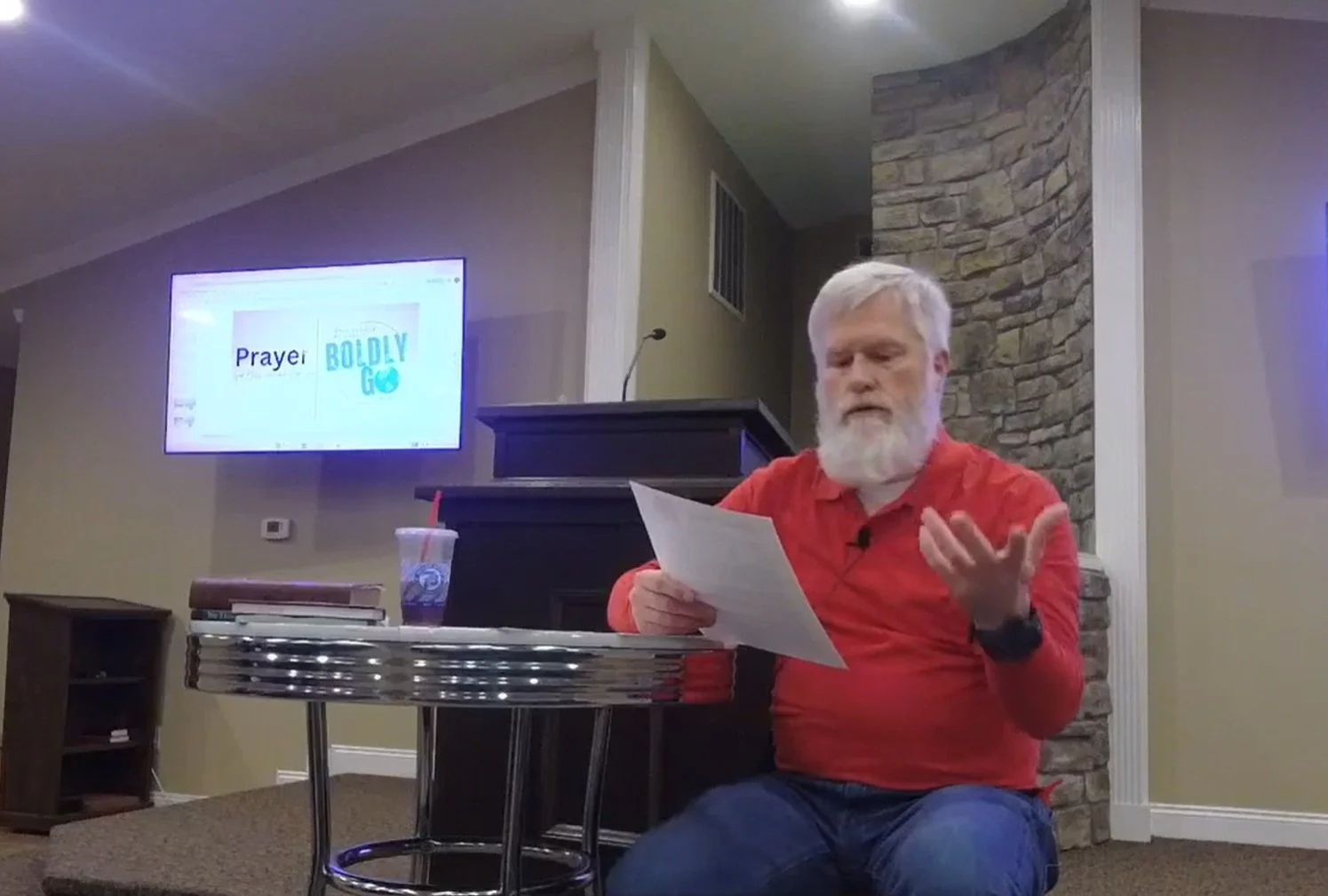 A man with a white beard and gray hair wearing a red shirt is sitting at a small round table, holding a piece of paper and gesturing with his hand. Behind him is a stone wall, a small podium with a microphone, and a screen displaying the words 'Prayer' and 'Boldly Go' in a church or community center setting.