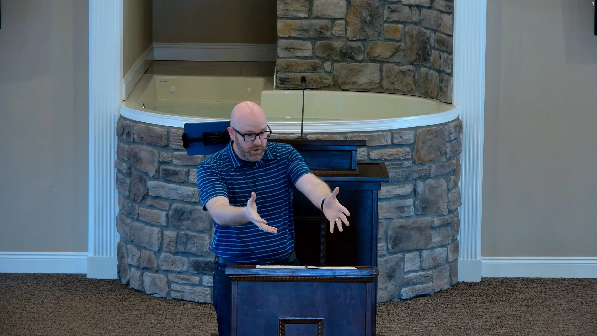 Bald man with glasses, wearing a blue striped polo shirt, speaking at a podium in front of a stone wall with a small hot tub in the background.