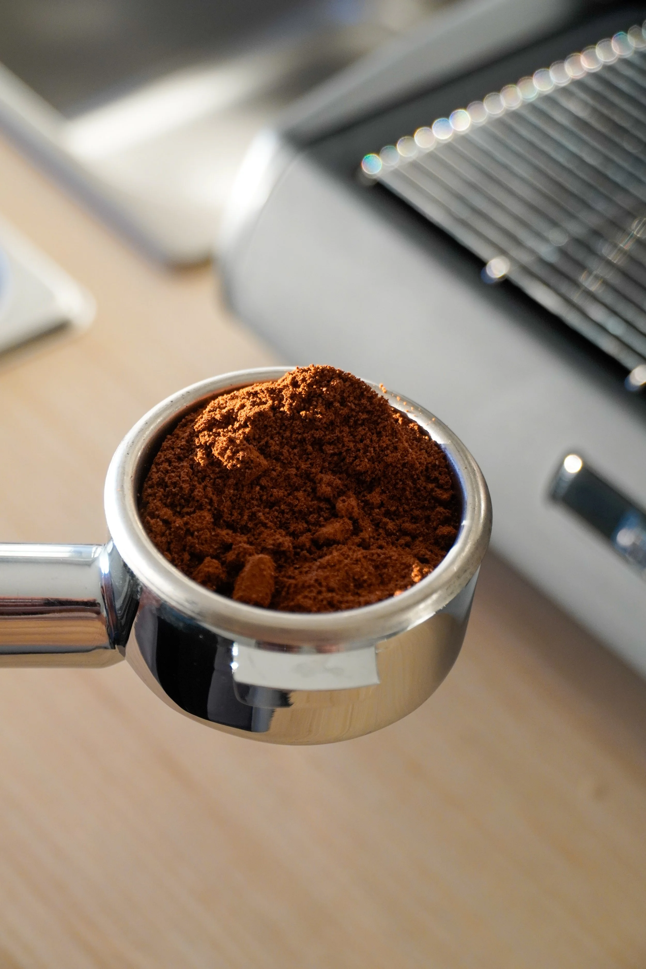 Close-up of a portafilter filled with finely ground coffee, held by a metal handle in front of a coffee machine.