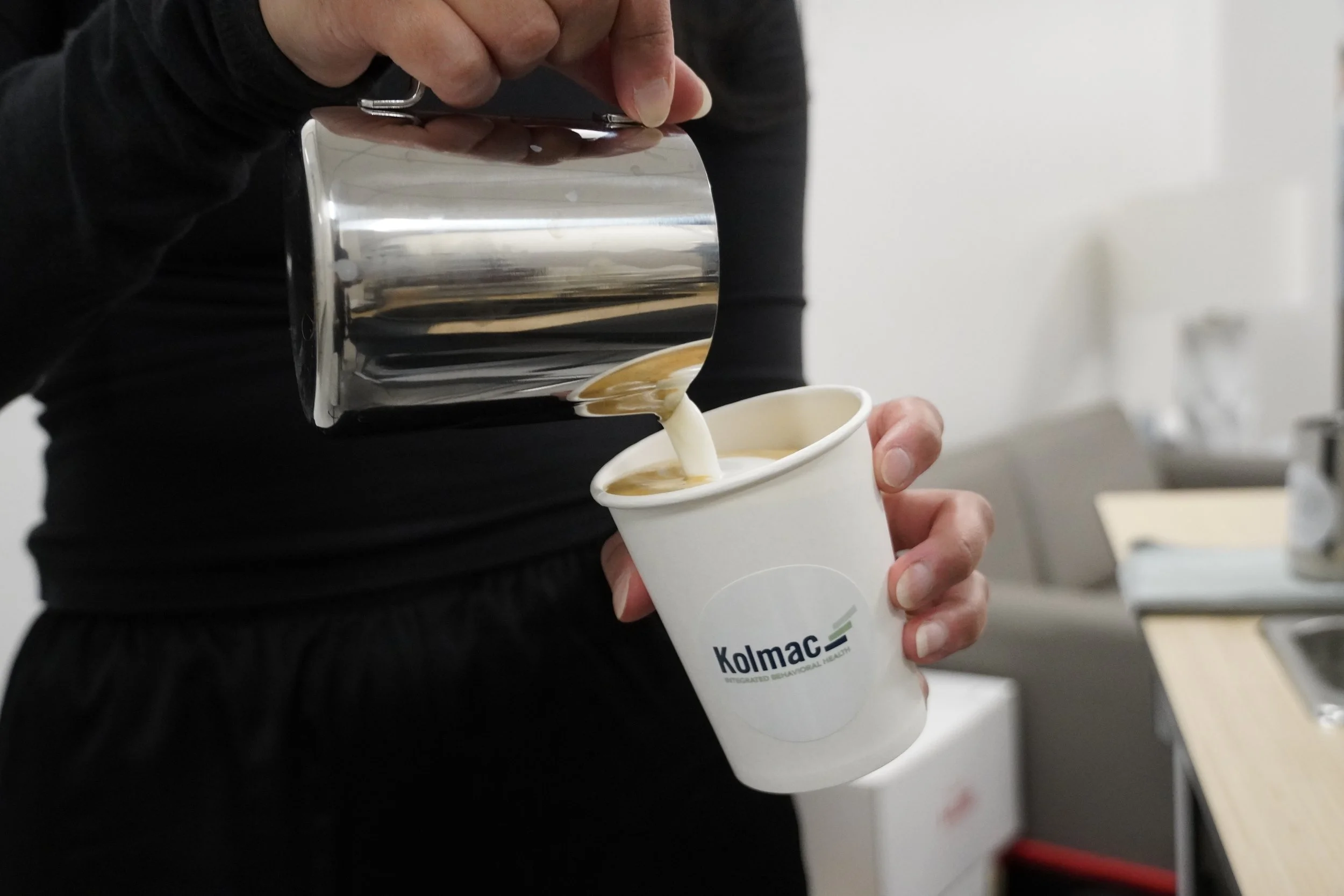 Person pouring a beverage into a white cup labeled 'Kolmac' from a metallic container.