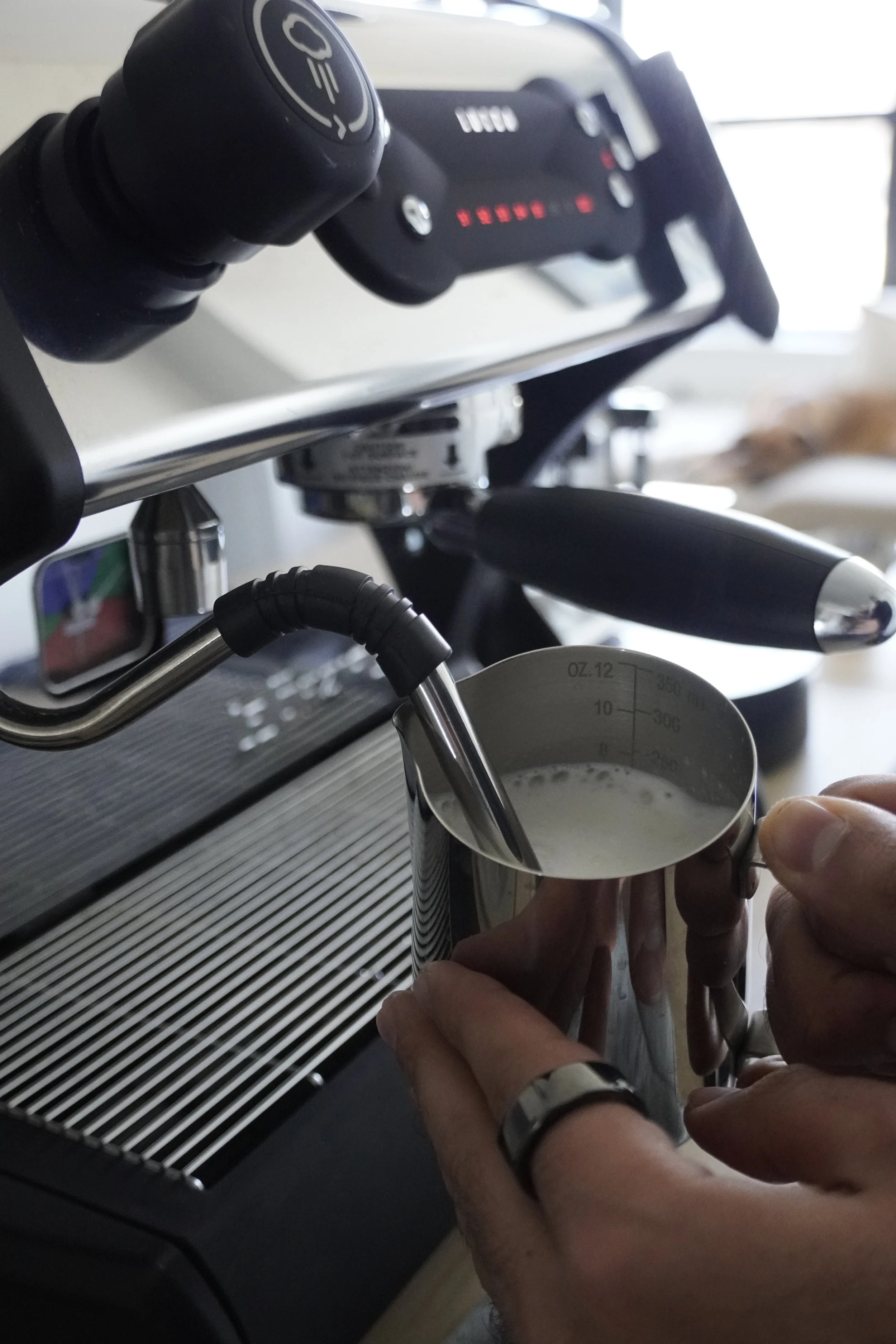 Hand holding a stainless steel milk frothing pitcher under an espresso machine's steam wand, creating milk foam.