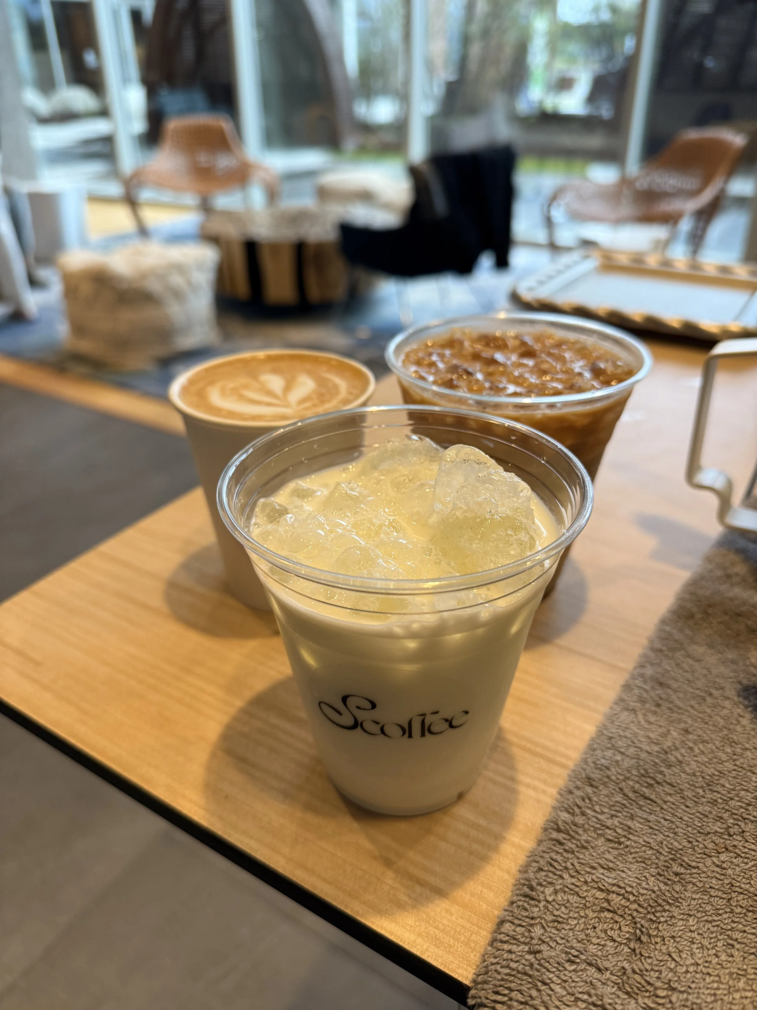 A latte with latte art, an iced matcha latte, and a cold brew coffee, in a cozy cafe setting with large windows and seating in the background.