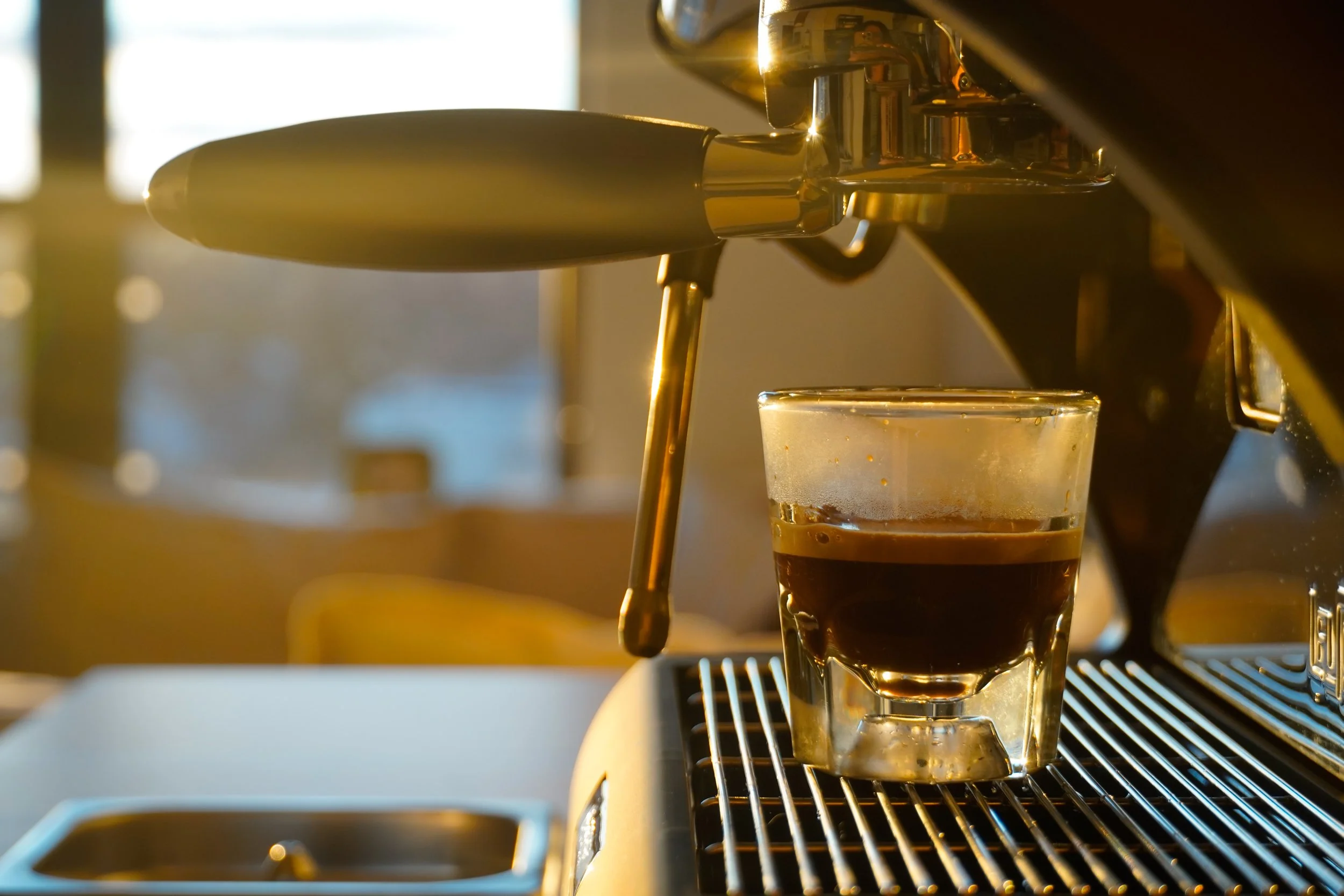 Close-up of a coffee machine pouring espresso into a glass cup, with warm sunlight in the background.