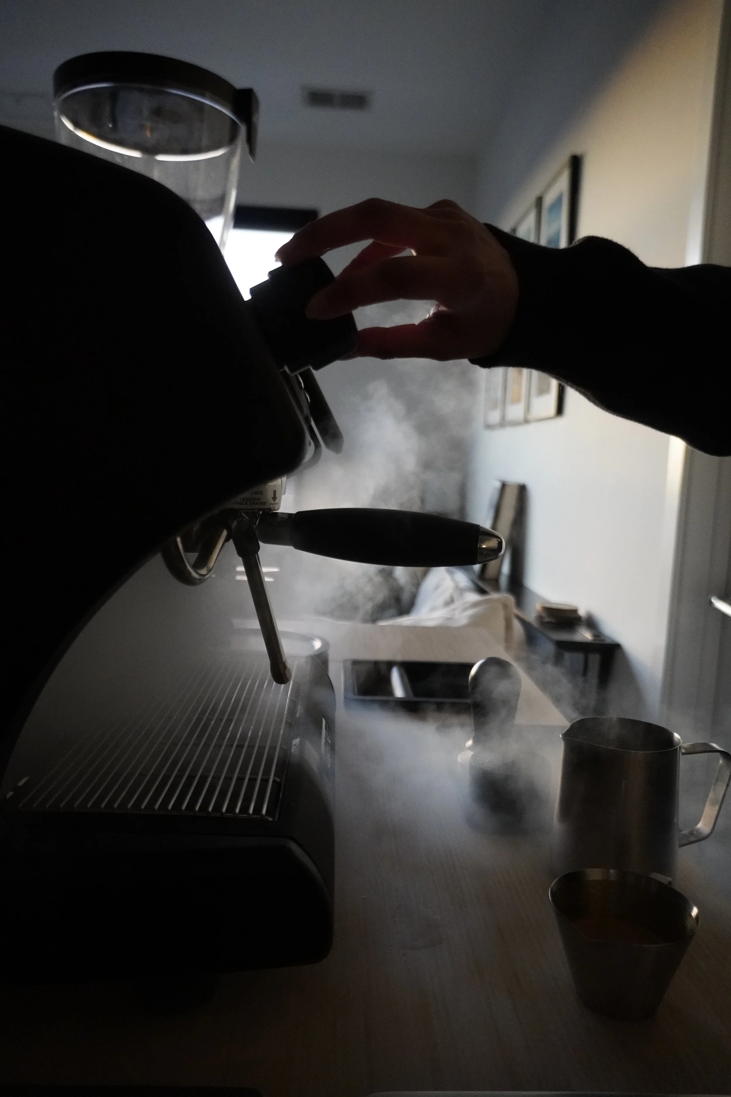 A person using a coffee machine, with steam rising as coffee brews, on a kitchen countertop with cups and a tablet nearby.