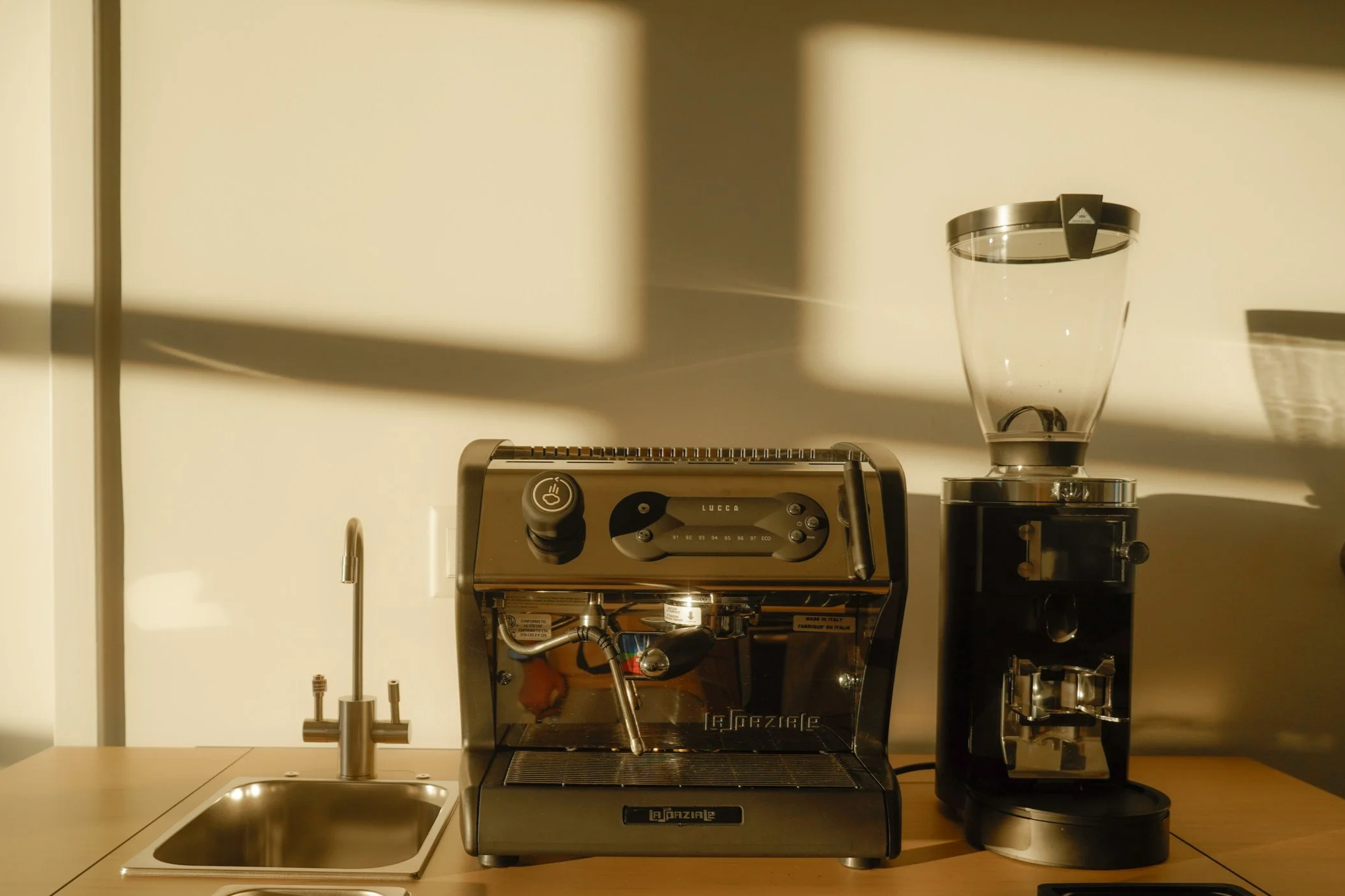Coffee machine, coffee grinder, and water faucet on a wooden counter in a kitchen with sunlight casting shadows on the wall.