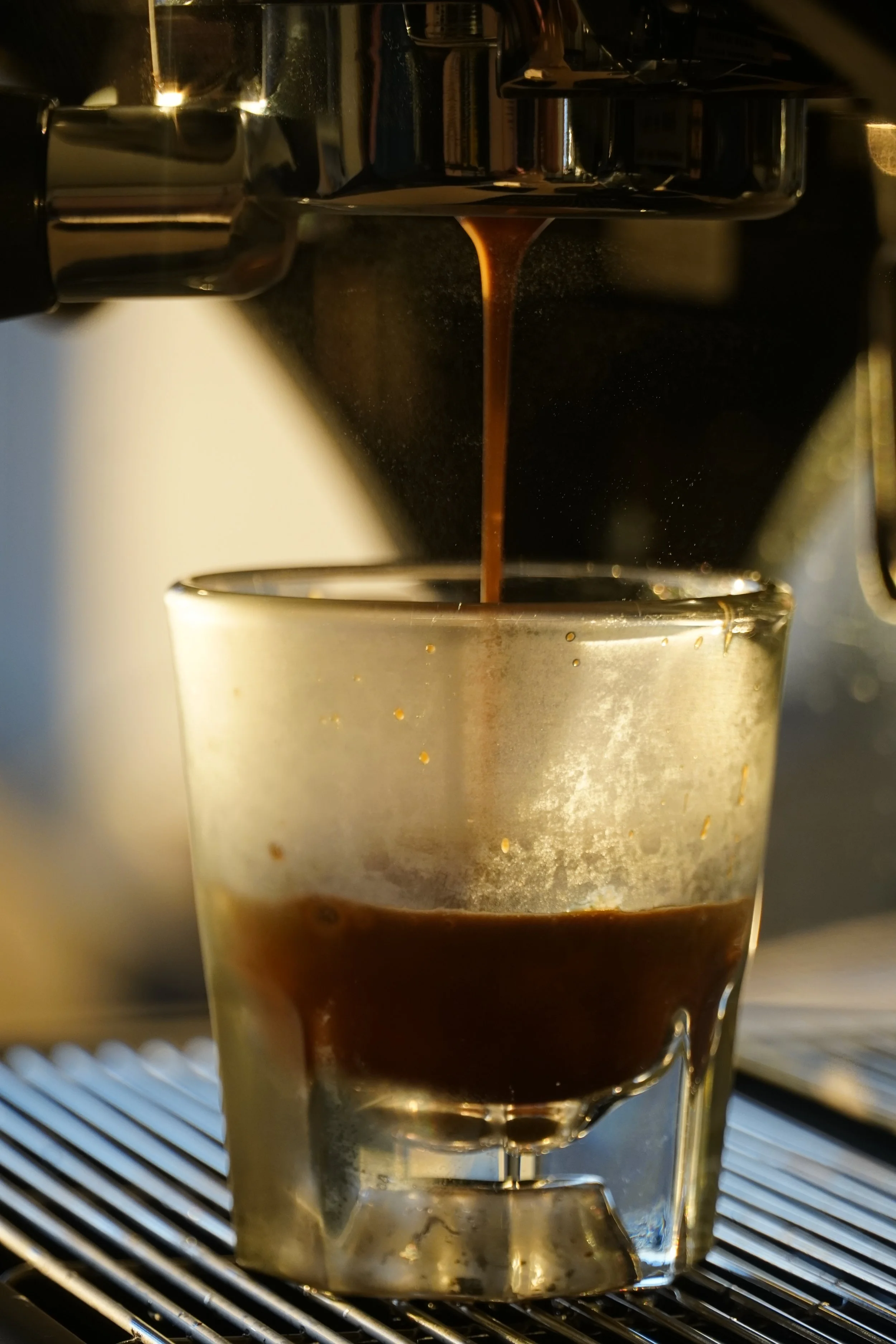 Close-up of hot espresso coffee being poured from an espresso machine into a glass cup with a metal container on top.