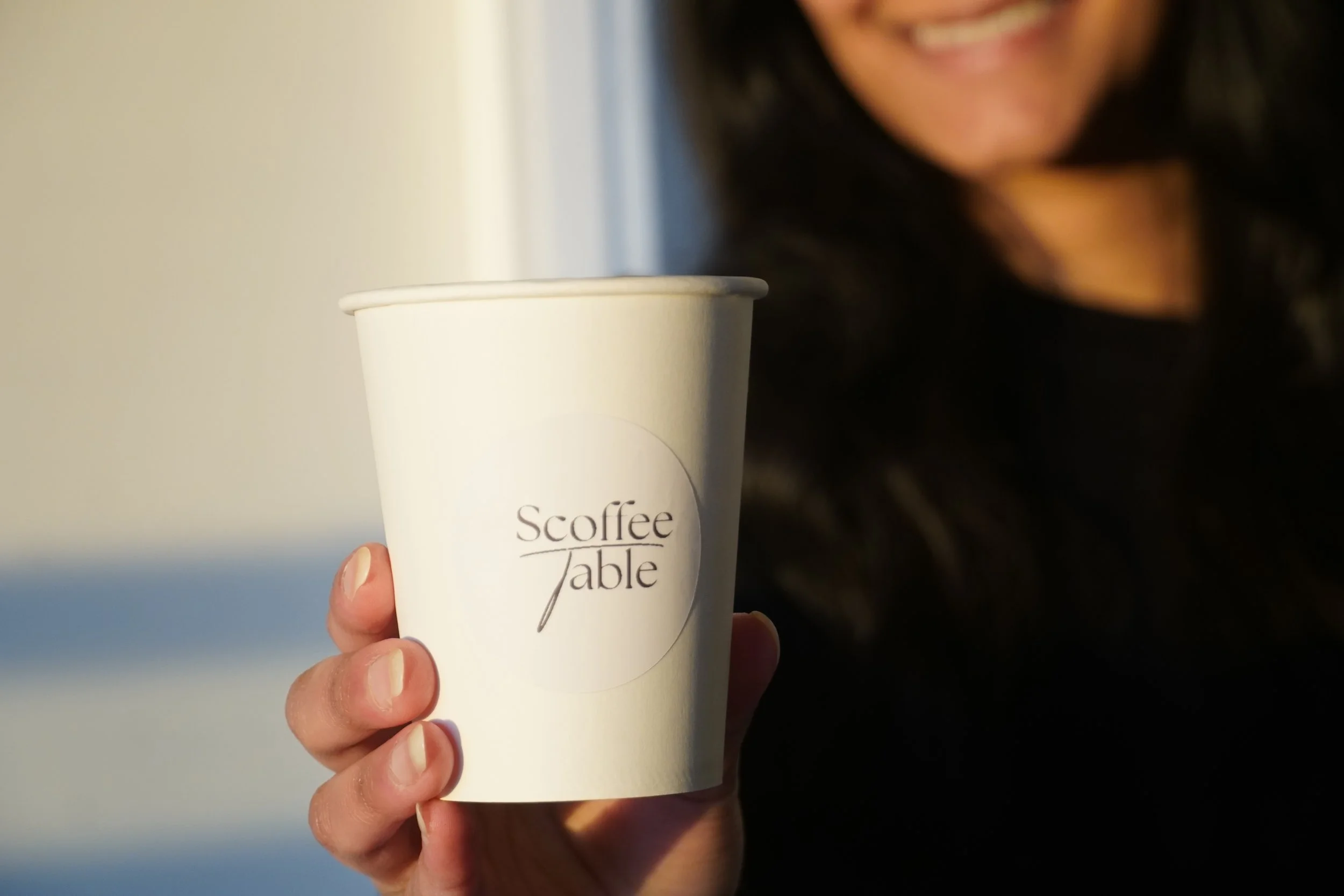 A person holding a white paper cup with the logo 'Scoffee Table' on it. The person's face is partially visible with a smile, and they have dark, curly hair. The background is blurred.