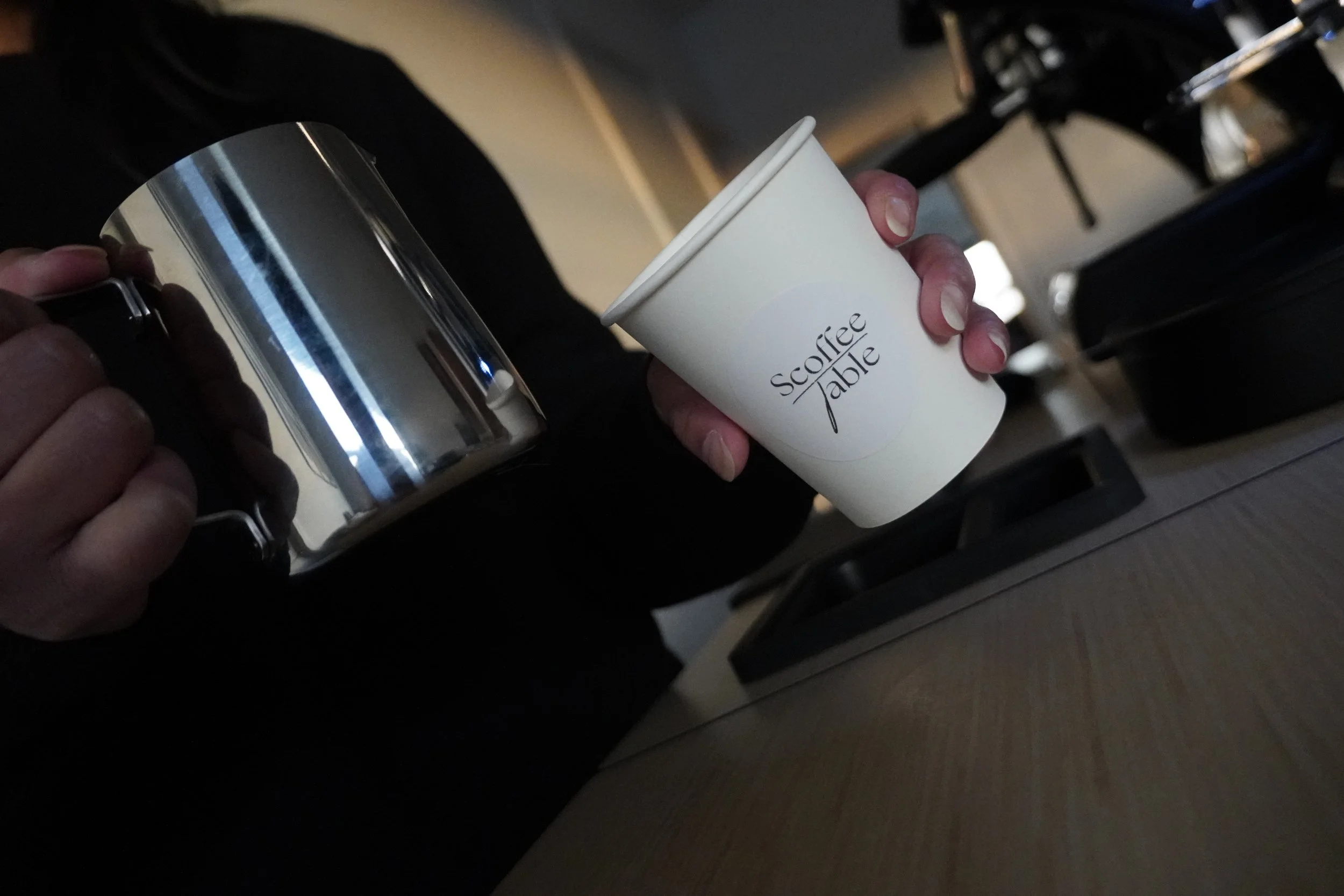 Person holding a stainless steel milk pitcher and a white paper coffee cup labeled "Scoffee Table" in a coffee shop or cafe setting.