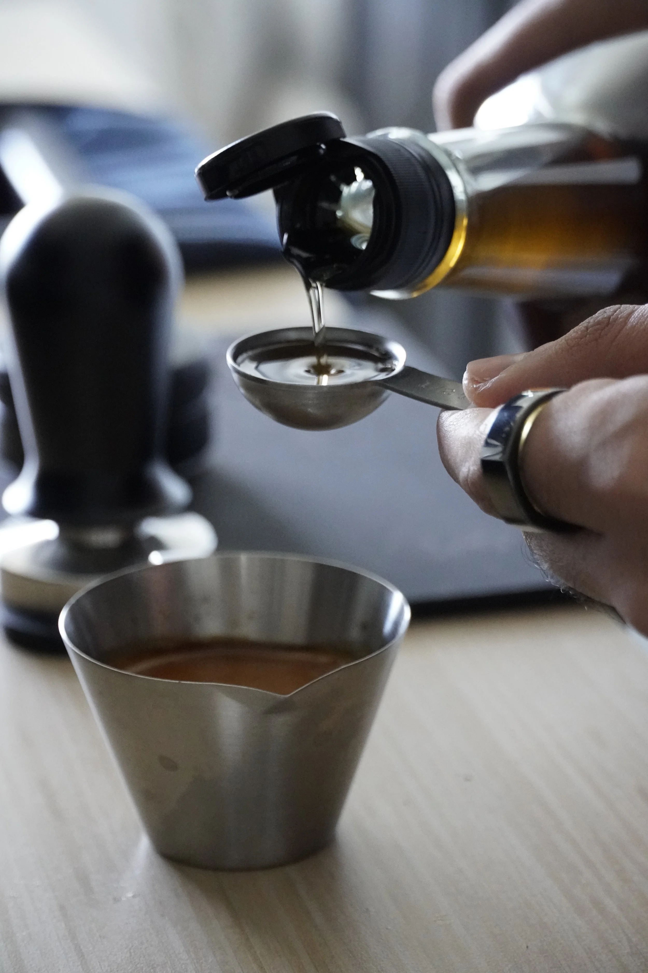 A person is pouring syrup from a bottle into a metal cup, with a measuring spoon.