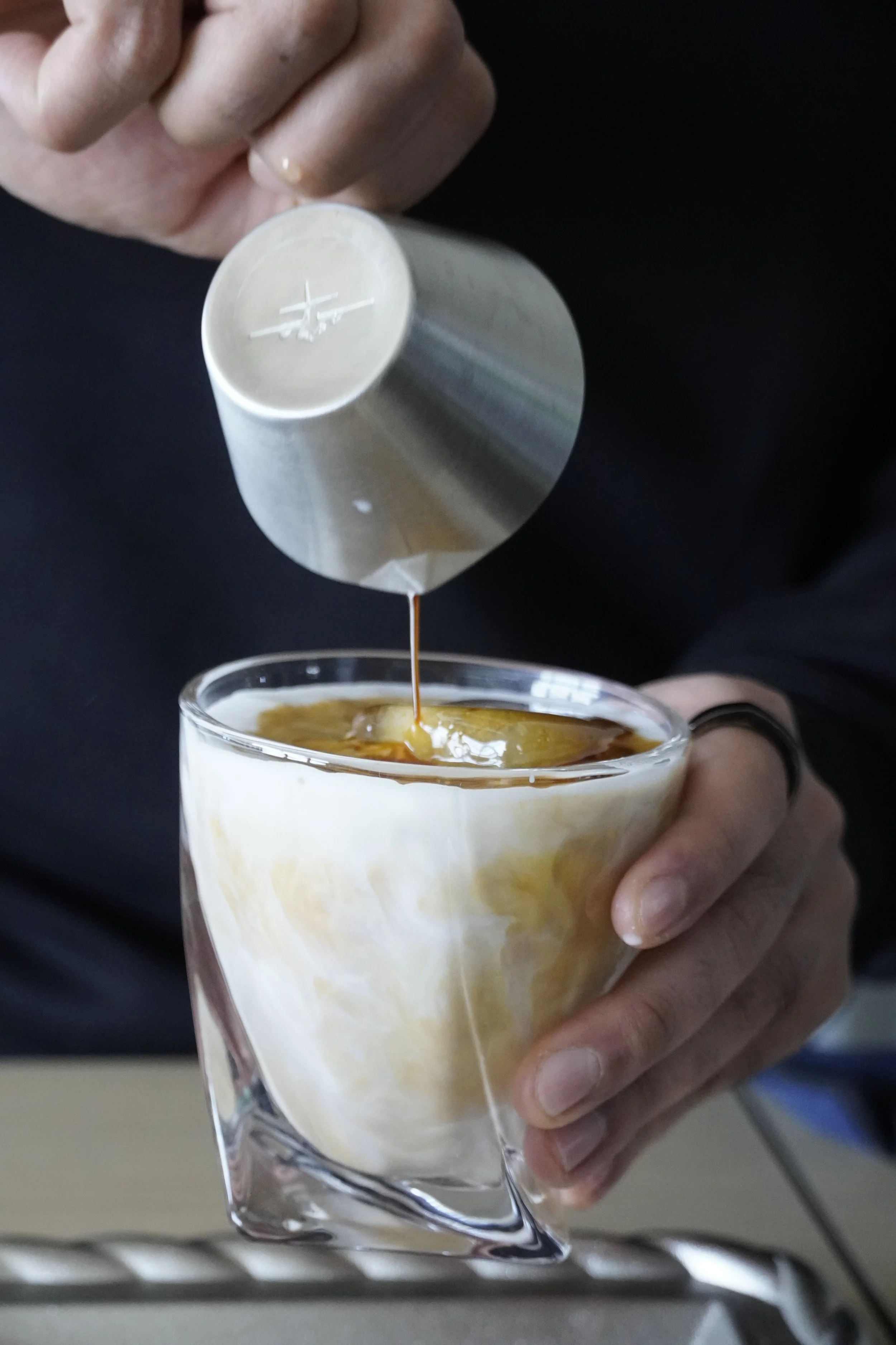 A person pouring coffee from a silver espresso cup into a glass cup of iced milk.