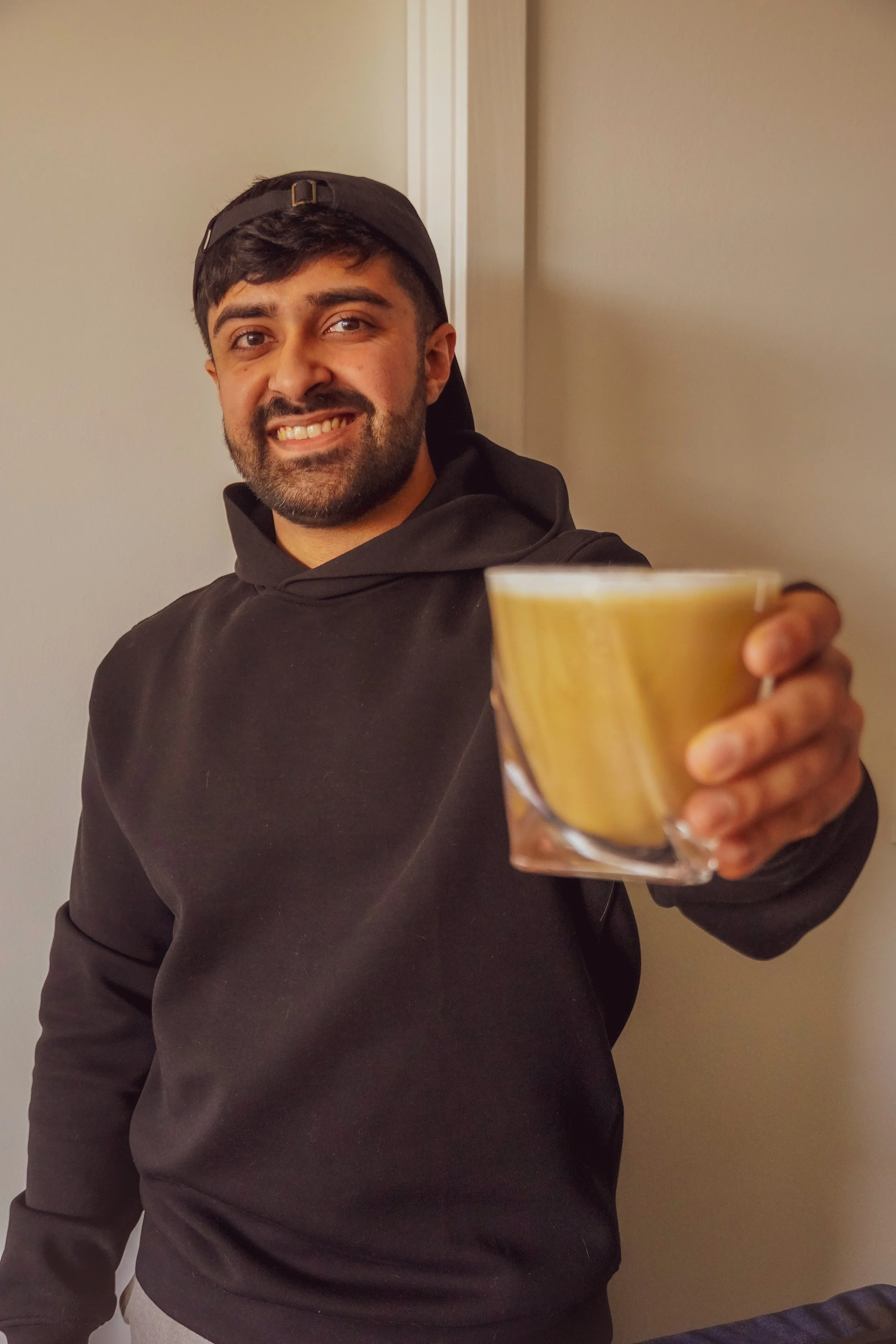 A man smiling and holding a cup of coffee in a glass mug towards the camera, standing indoors in front of a plain wall.