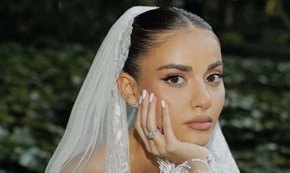 Close-up portrait of a woman wearing a wedding veil, with makeup and jewelry, resting her chin on her hand.