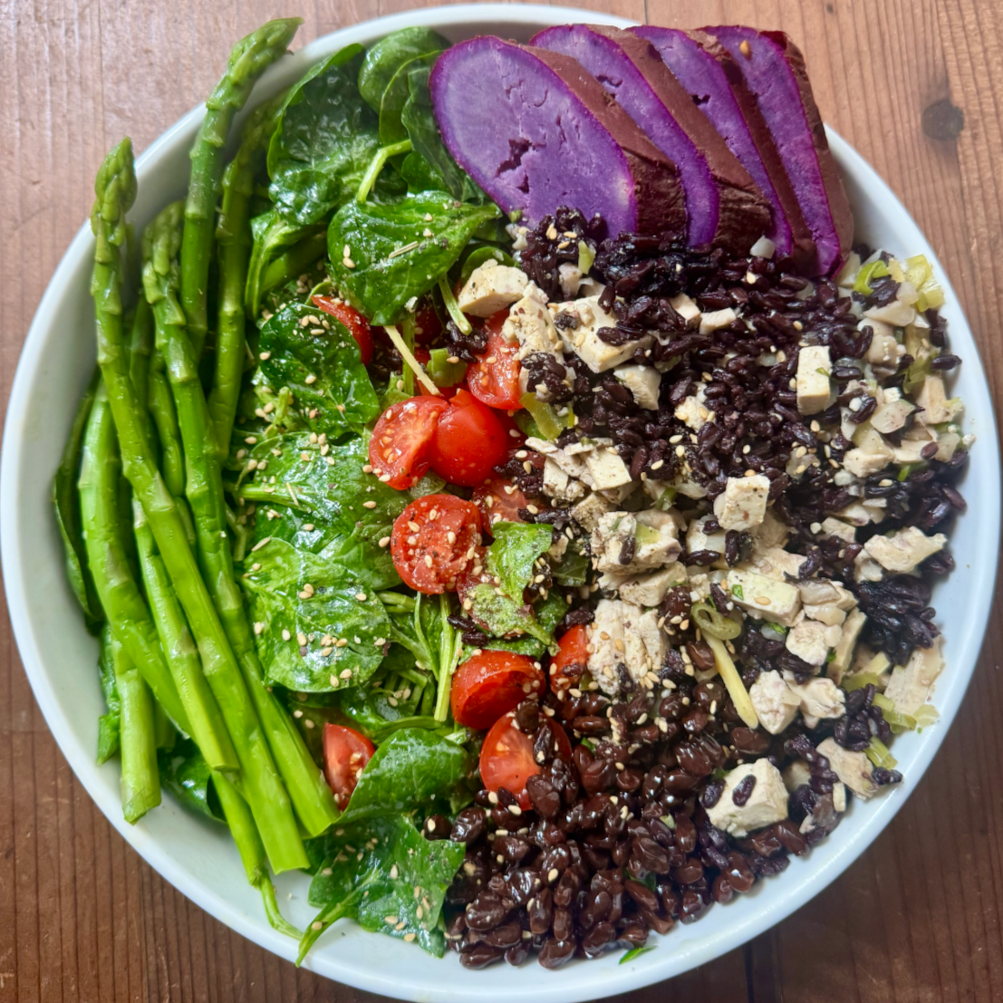 Bowl of mixed salad with asparagus, spinach, cherry tomatoes, purple sweet potatoes, shredded chicken, black beans, and sesame seeds on a wooden surface.