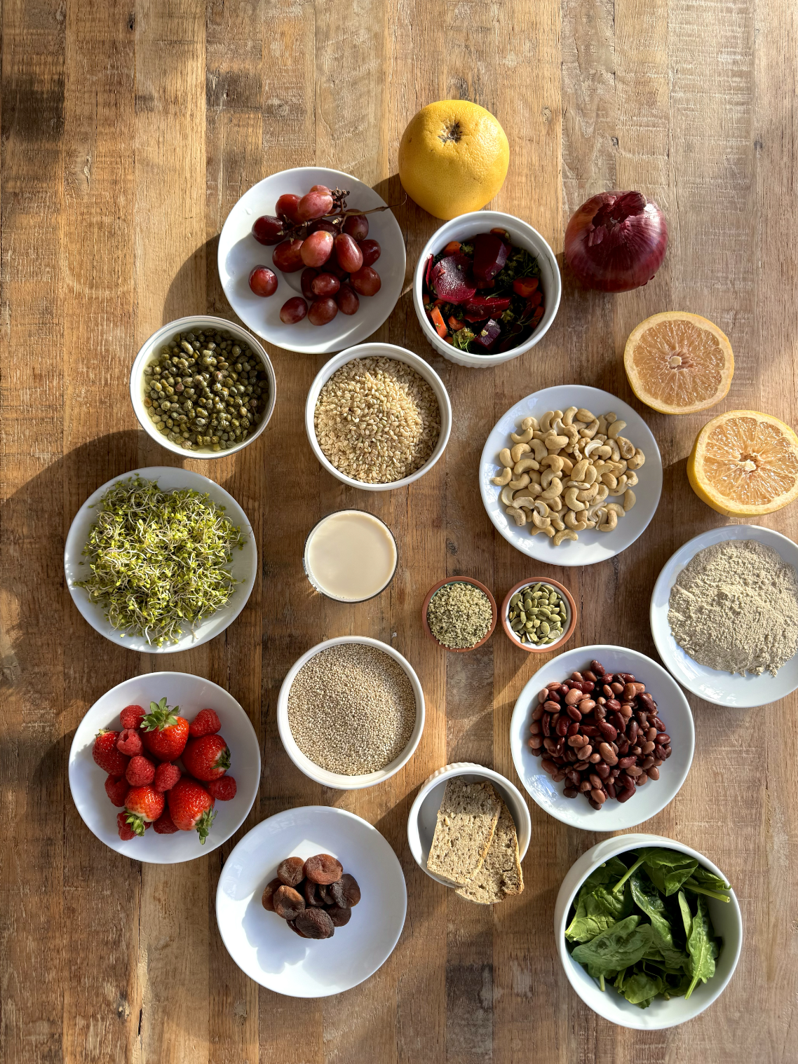 An assortment of fresh fruits, vegetables, grains, nuts, seeds, and greens arranged on a wooden table, including grapes, grapefruit, strawberries, dried dates, cashews, lentils, spinach, and various seasonings.