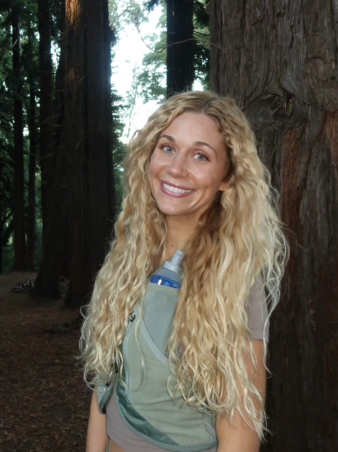 Smiling woman with curly blonde hair in a forest, wearing a light-colored sleeveless shirt and a small green backpack with a water bottle.
