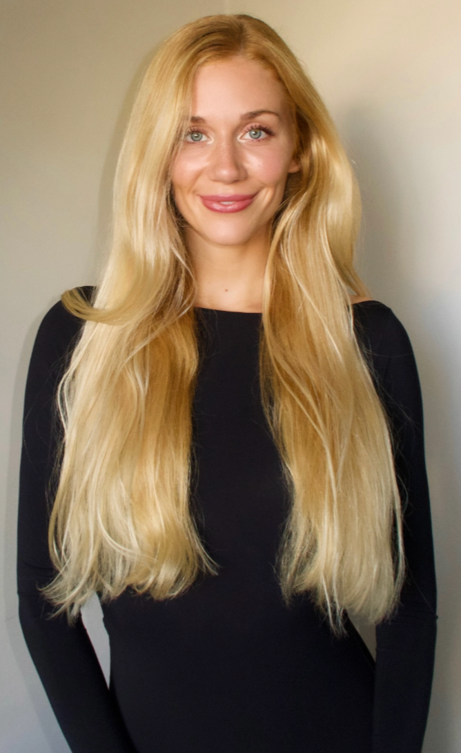 A woman with long, wavy blonde hair, wearing a black top, smiling against a plain background.