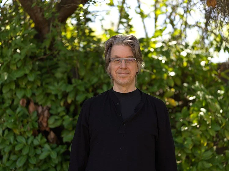 A man with brown and gray hair and glasses stands outdoors in front of green leafy trees, wearing a black shirt and smiling.