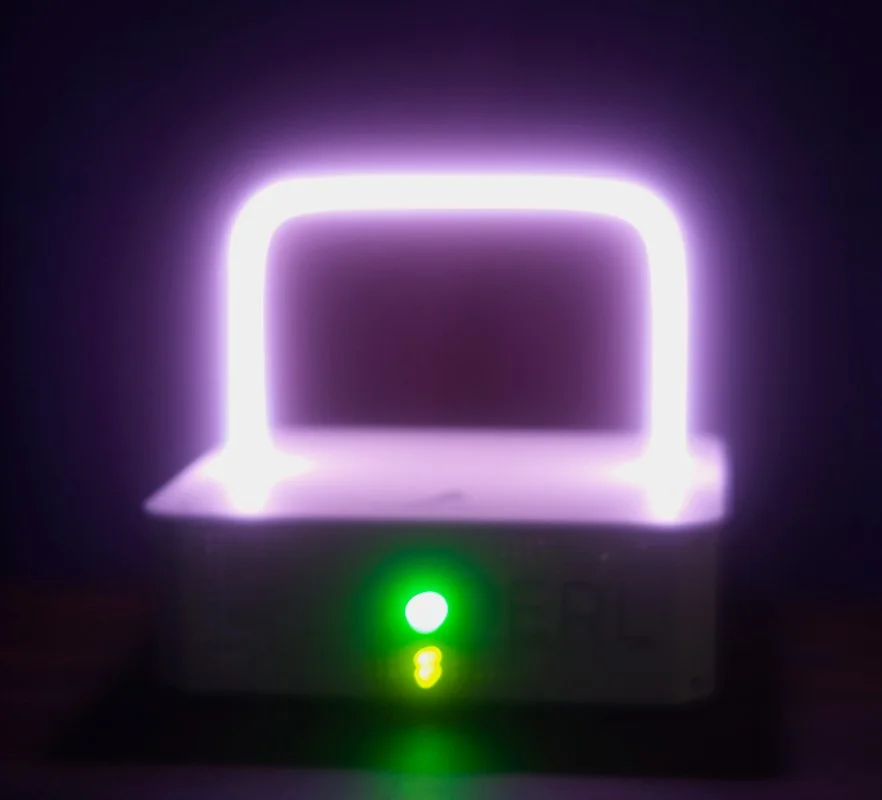 A PERL Rife device surrounded in darkness, with a rectangular violet plasma glowing in the dark above a glowing green indicator. The image is glowing more than the other PERL Rife image, as if it is melting down.