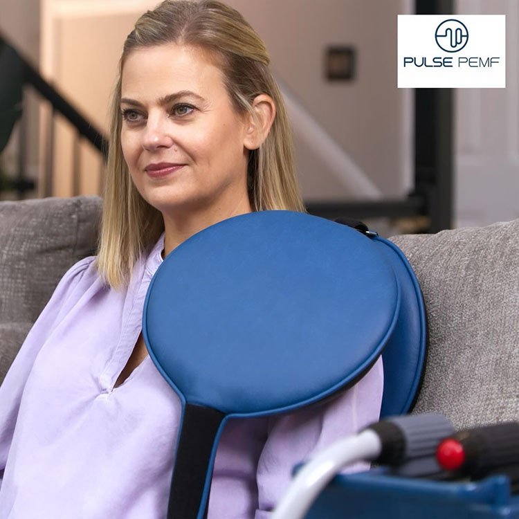 A woman with blue Pulse paddles sandwiched in front and back of her shoulder receives PEMF treatment from a Pulse XL Pro, which sits beside her.