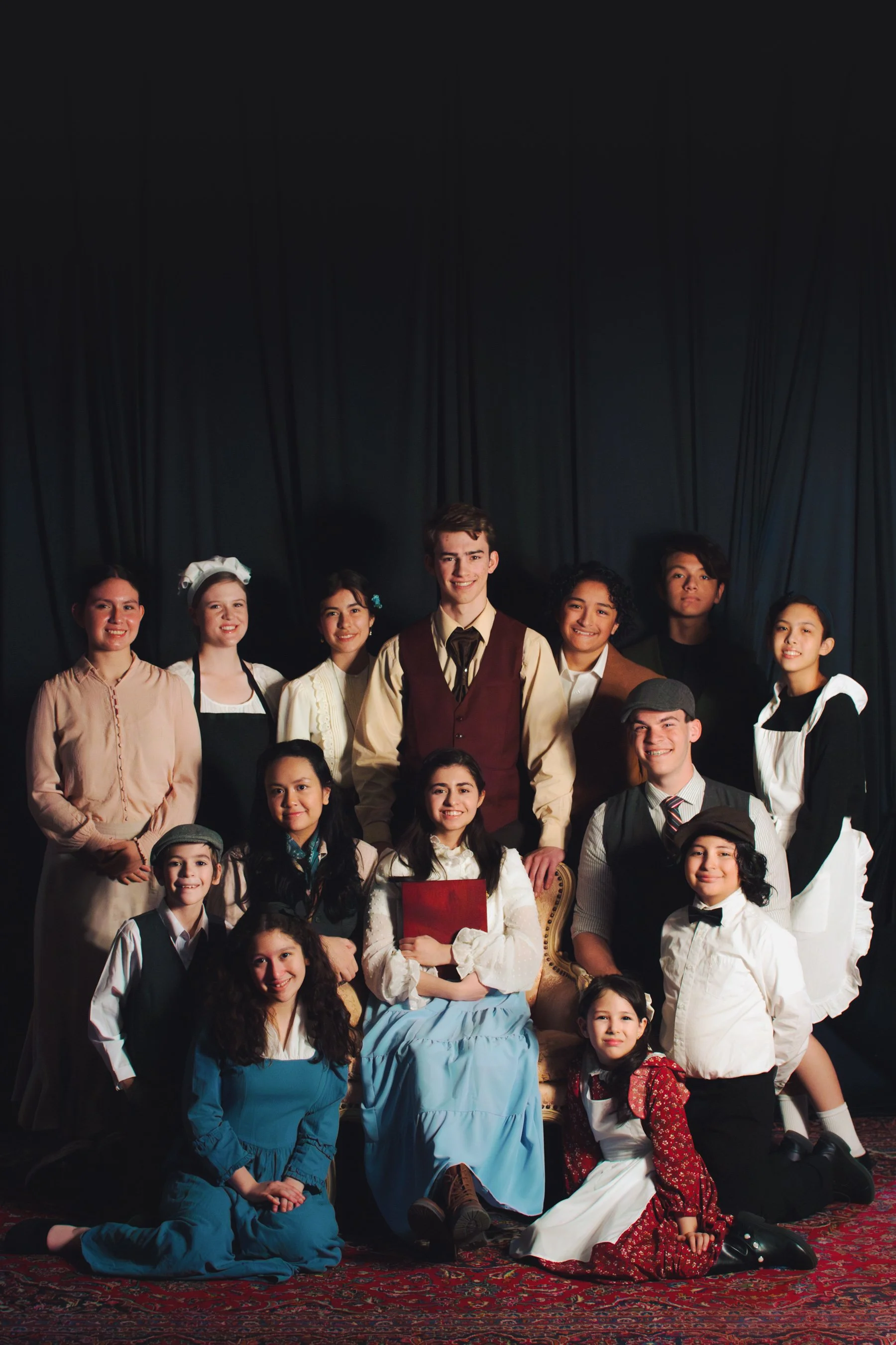 A group of young actors dressed in period costumes on stage, posing for a photo during a theater production.