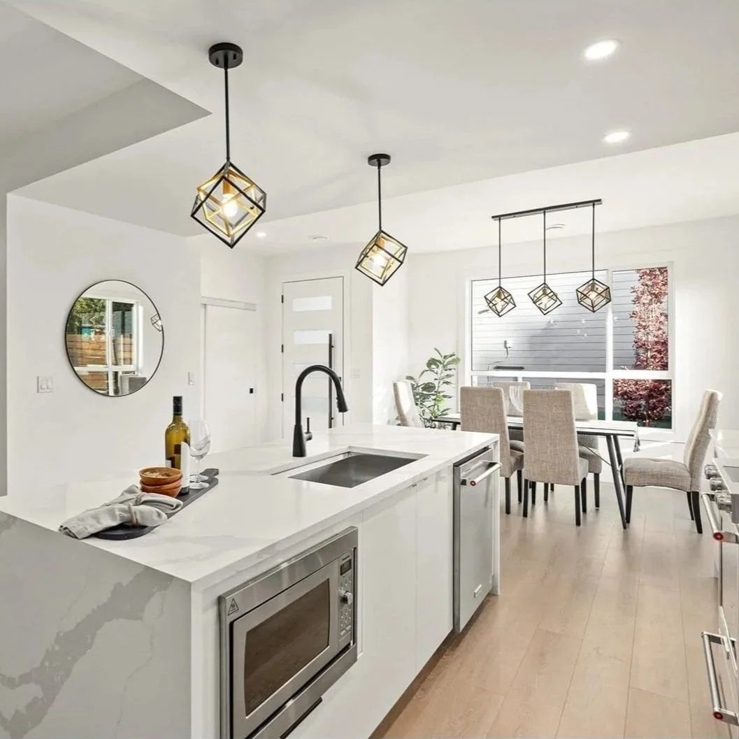 Modern kitchen with white countertops, black faucet, and built-in microwave, with dining area featuring a dark table and beige chairs, pendant or chandelier lighting, and large windows bringing in natural light