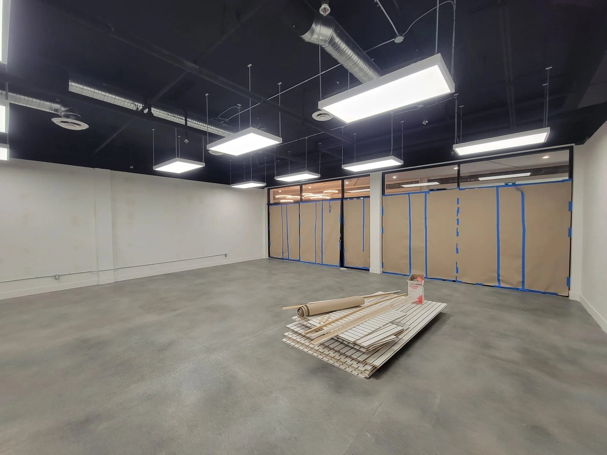 Interior of a room under construction with black ceiling, white walls, suspended rectangular ceiling lights, ductwork, and construction materials on the floor.