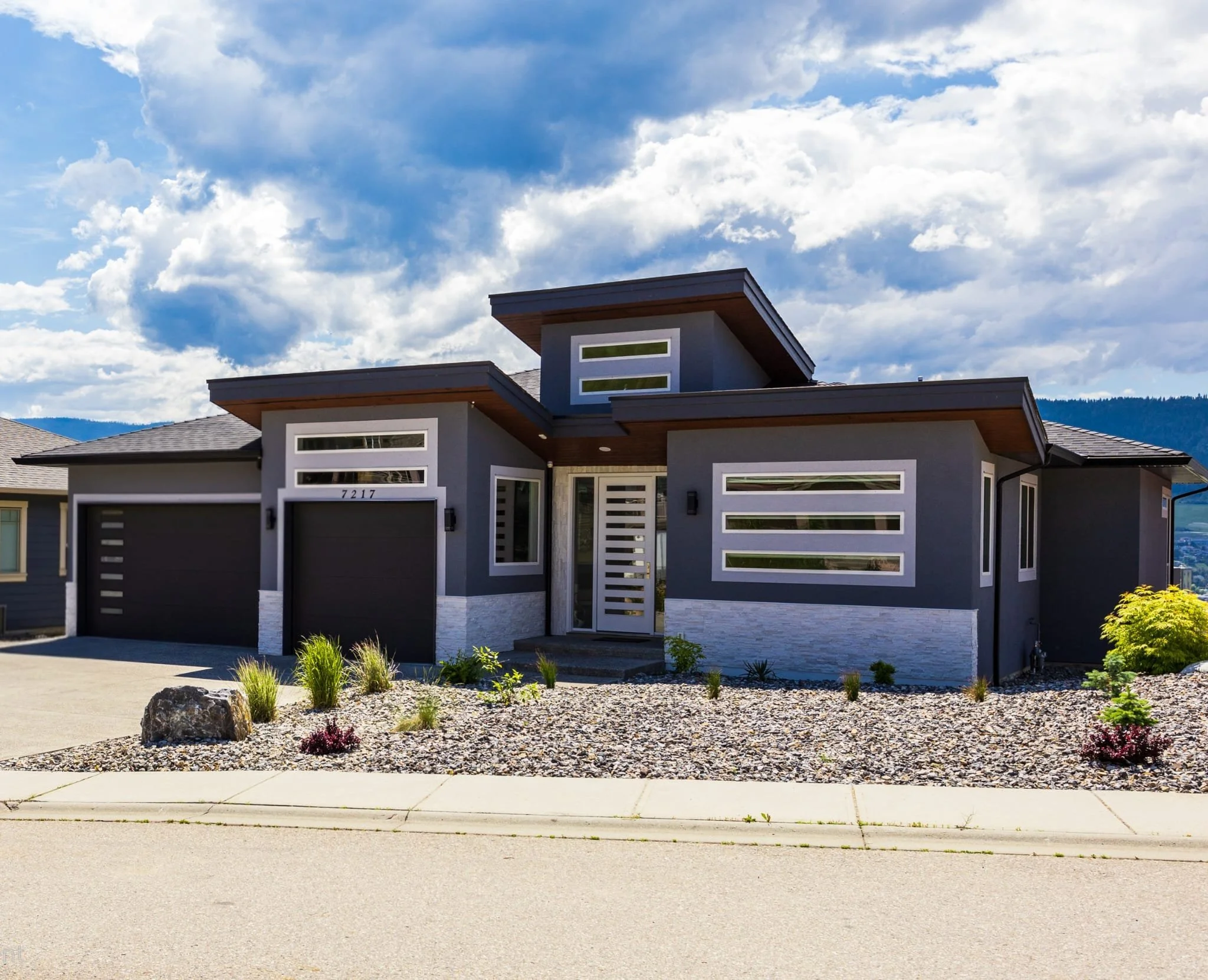 Modern house with flat roof, gray walls, and large windows, front yard with rocks, plants, and a concrete driveway, blue sky with scattered clouds in the background.