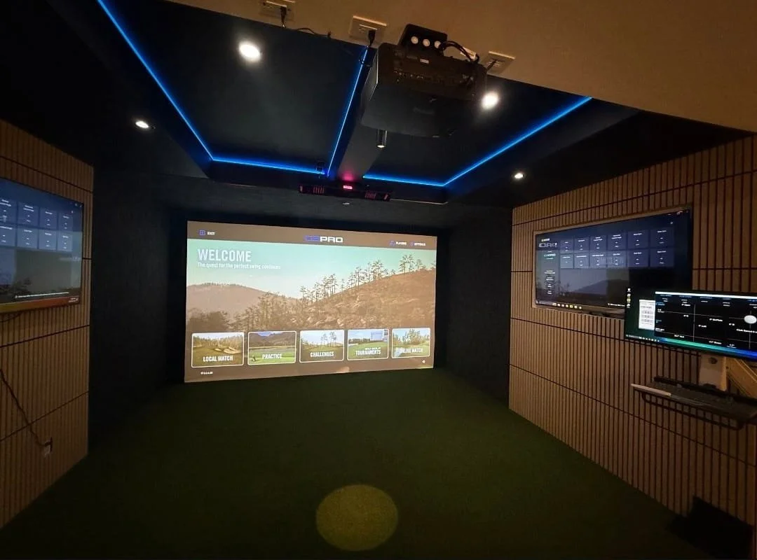 Indoor golf simulation room with a large screen displaying a welcome message and various game options, surrounded by multiple monitors and wood-paneled walls with blue ambient lighting.