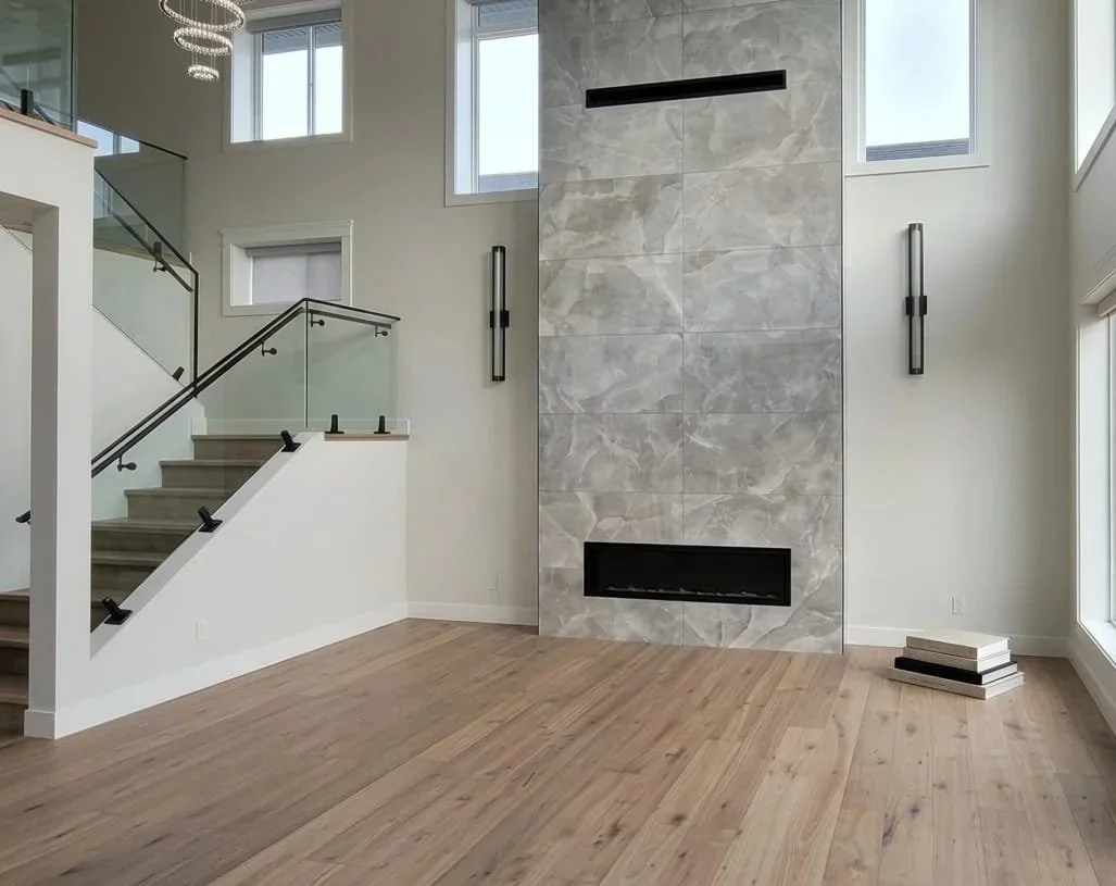Modern living room with a tall gray stone fireplace, hardwood floors, large windows, and a staircase with glass railing.
