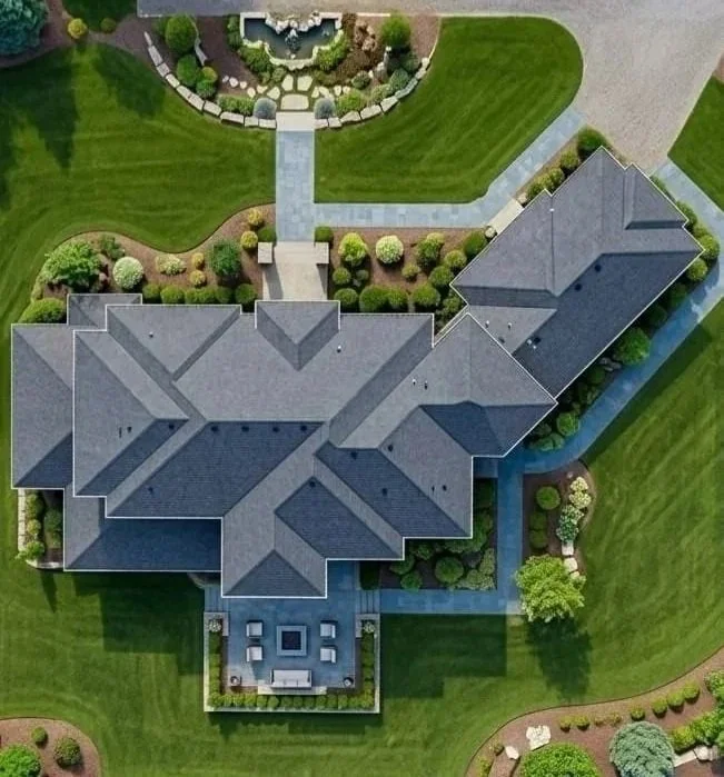 Aerial view of a large house with a complex roof design, surrounded by a well-maintained lawn, pathways, and landscaped gardens with trees and shrubs.