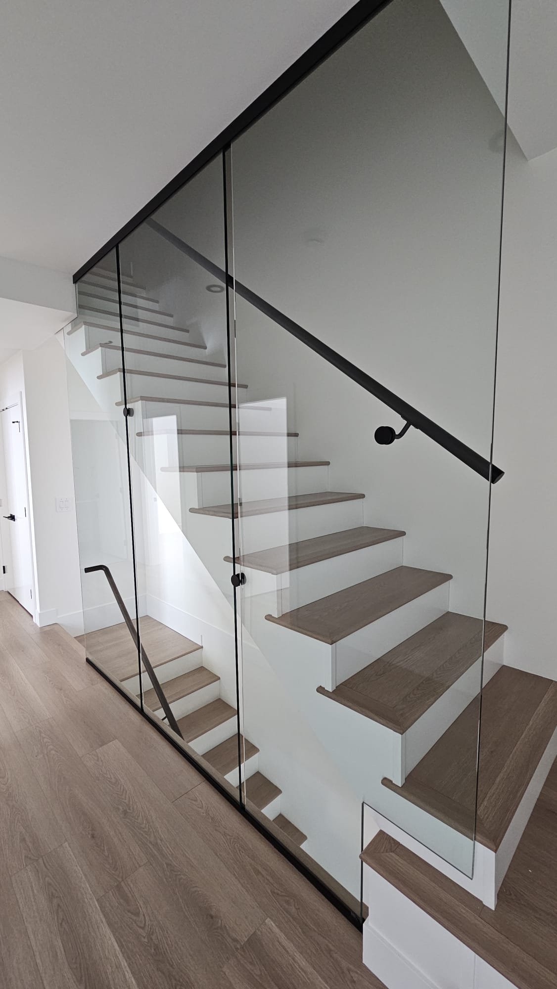 A modern staircase with wooden steps and a glass railing, viewed from a lower angle in a minimalist, well-lit interior.